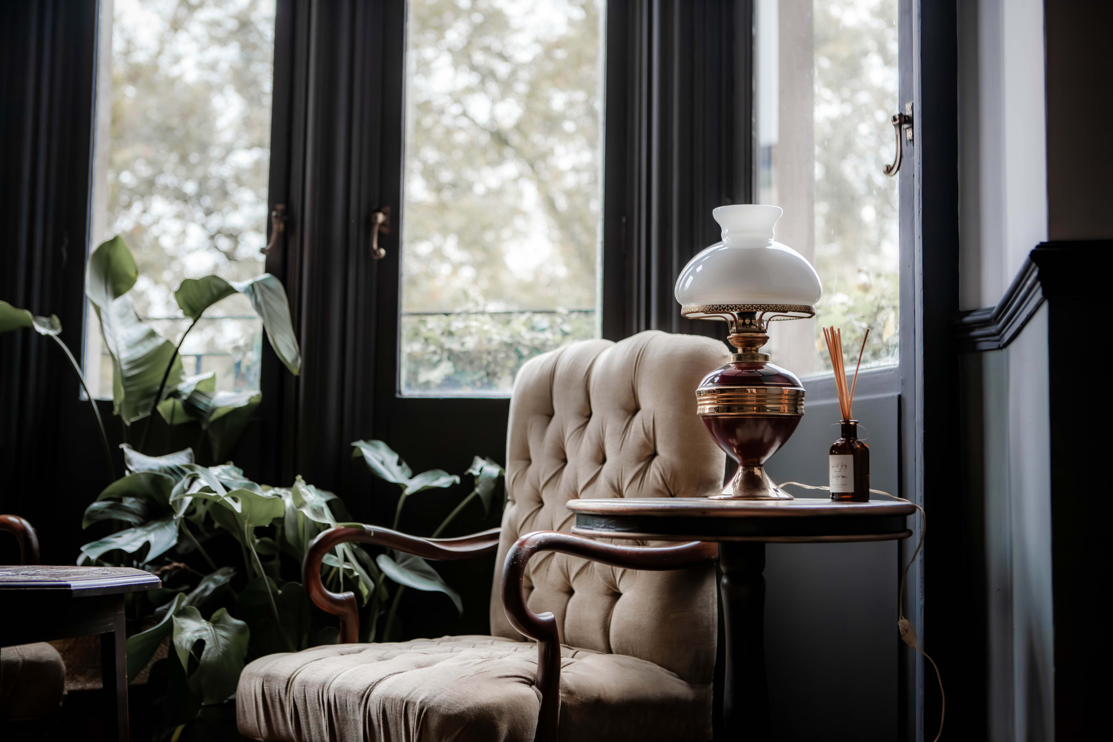 A tufted armchair is positioned next to a small table with a vintage lamp and a decorative bottle, surrounded by large plants and sunlight filtering through dark windows.