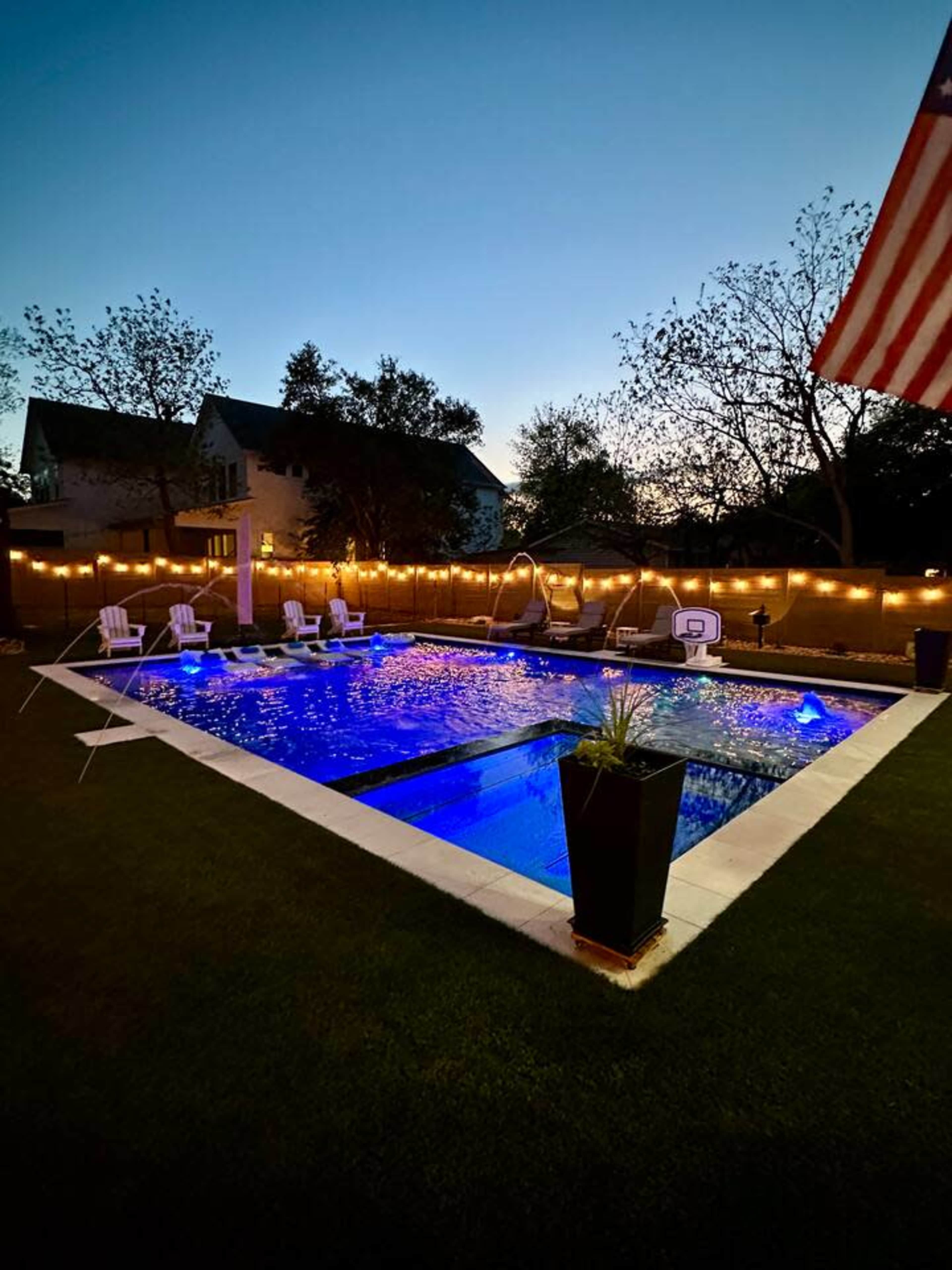 The image shows a well-lit backyard swimming pool surrounded by lawn chairs and string lights at dusk.