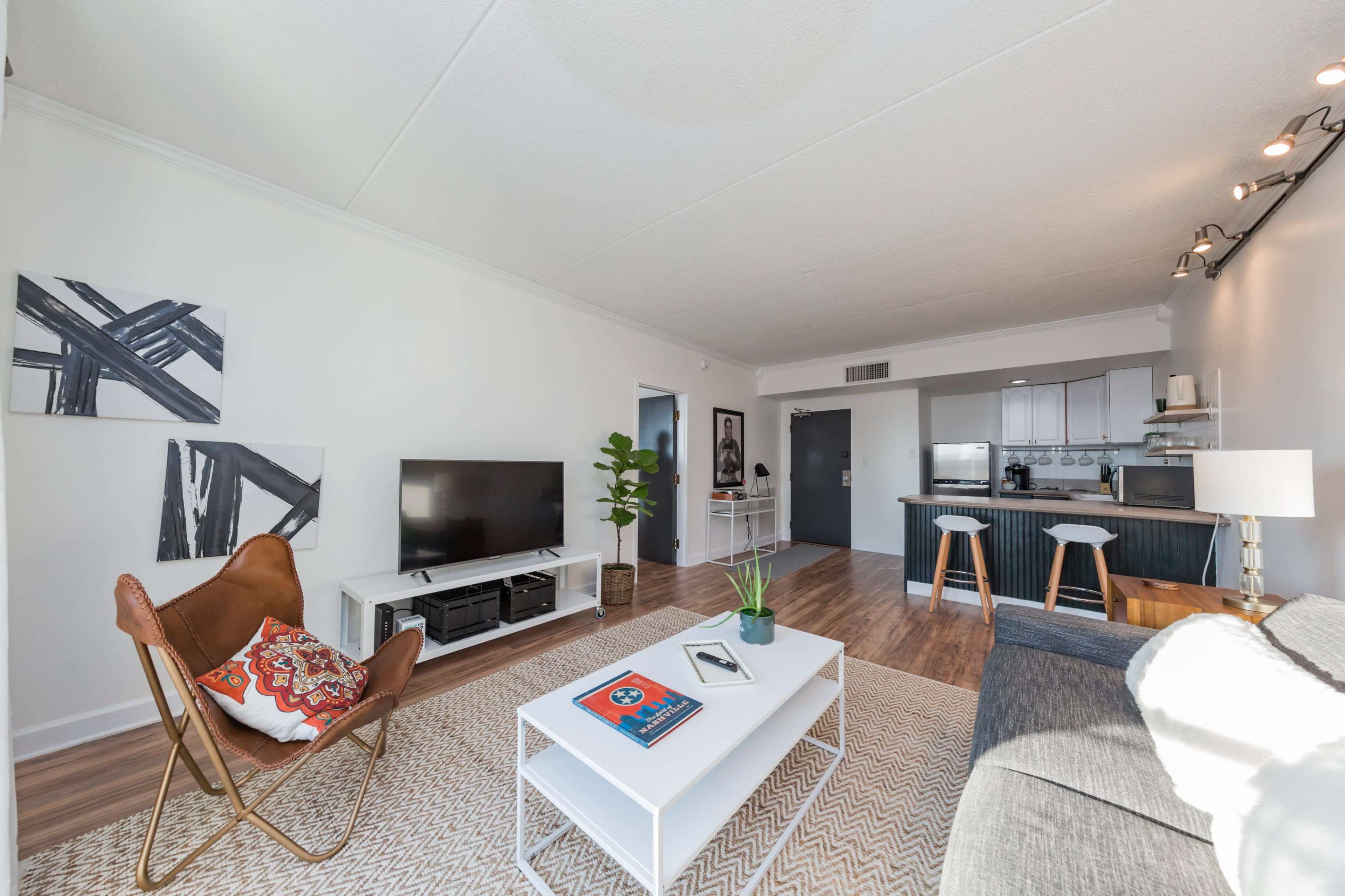 A modern living room with a television, a coffee table, and a combination of seating options including a chair and a sofa, leading into a kitchen area in the background.