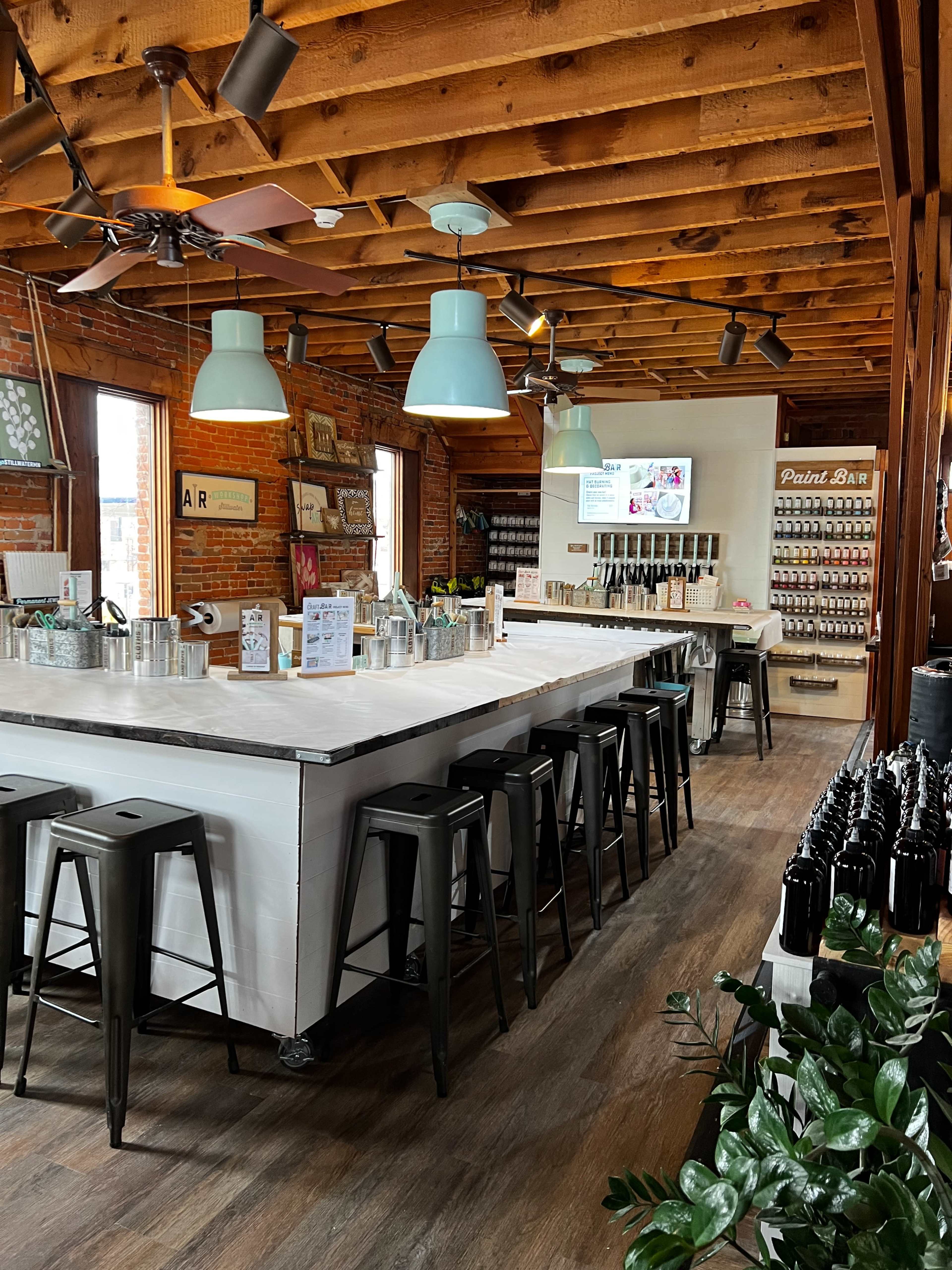 The image shows a spacious interior of a craft brewery or art studio with a large white bar area, metal stools, and wooden beams.