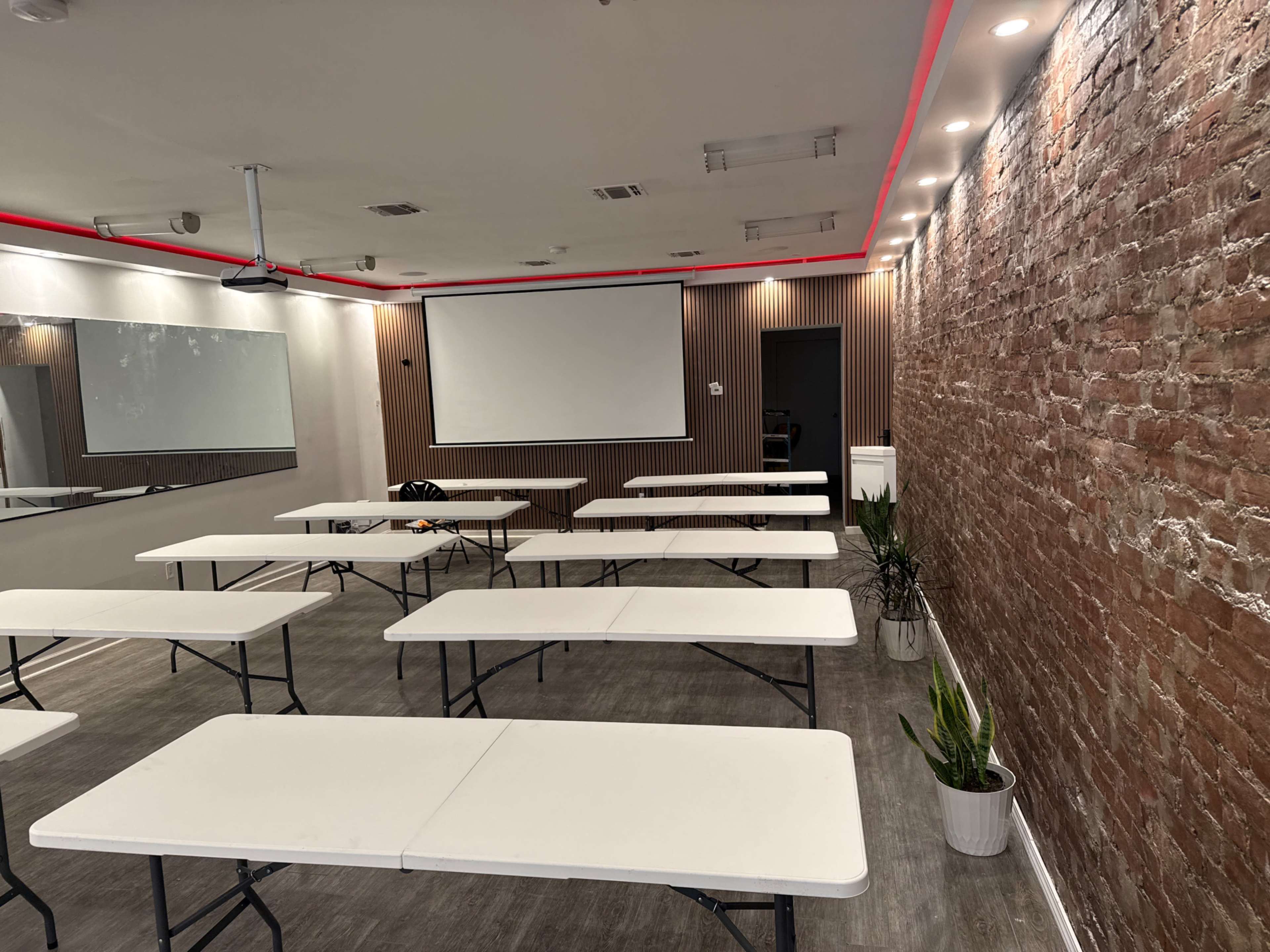The image shows a modern meeting room featuring several folding tables arranged in rows, with a large screen and a brick accent wall.