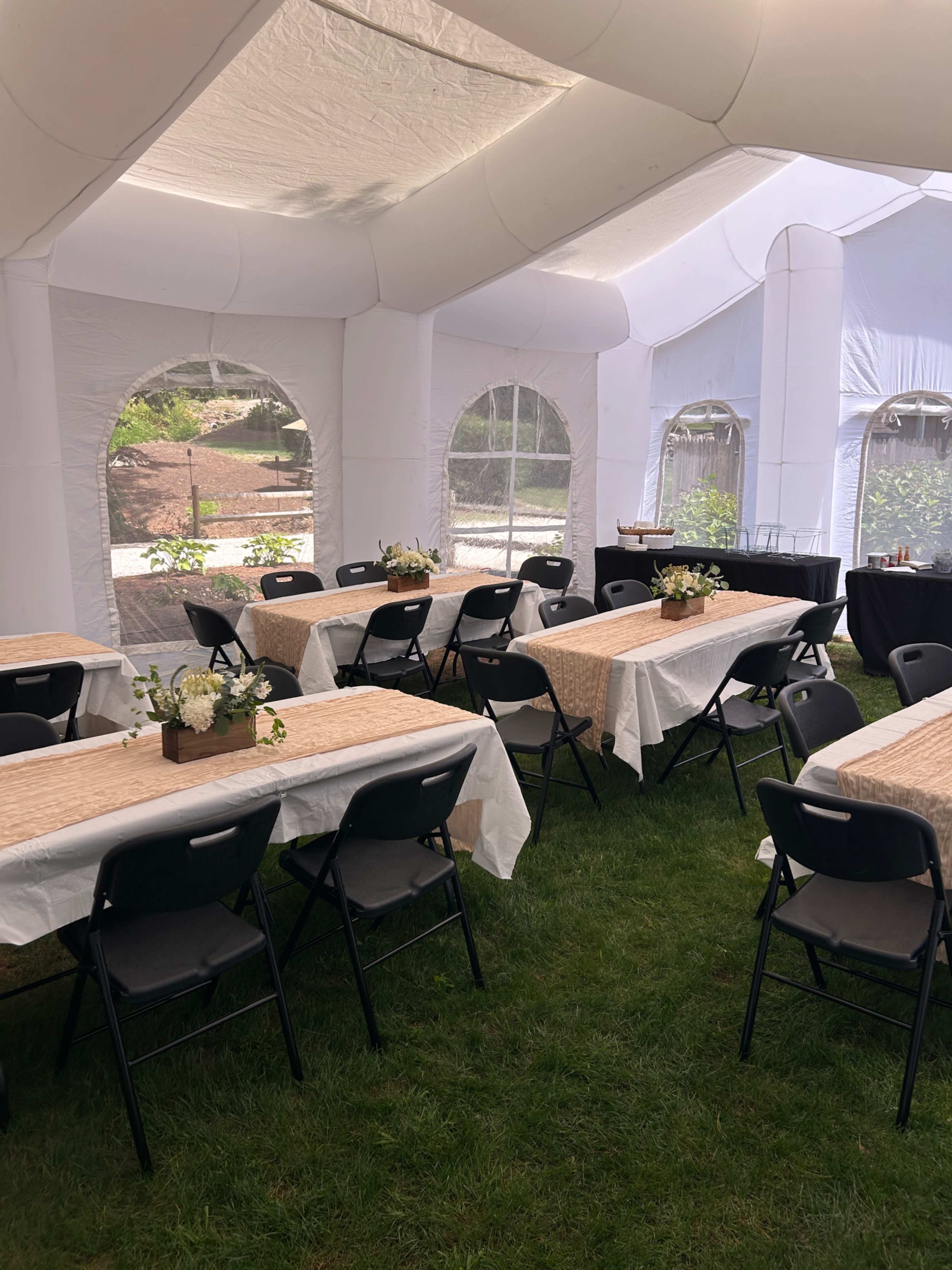 The image shows a large white tent set up with several tables decorated with table runners and floral centerpieces, all arranged on a grassy area.