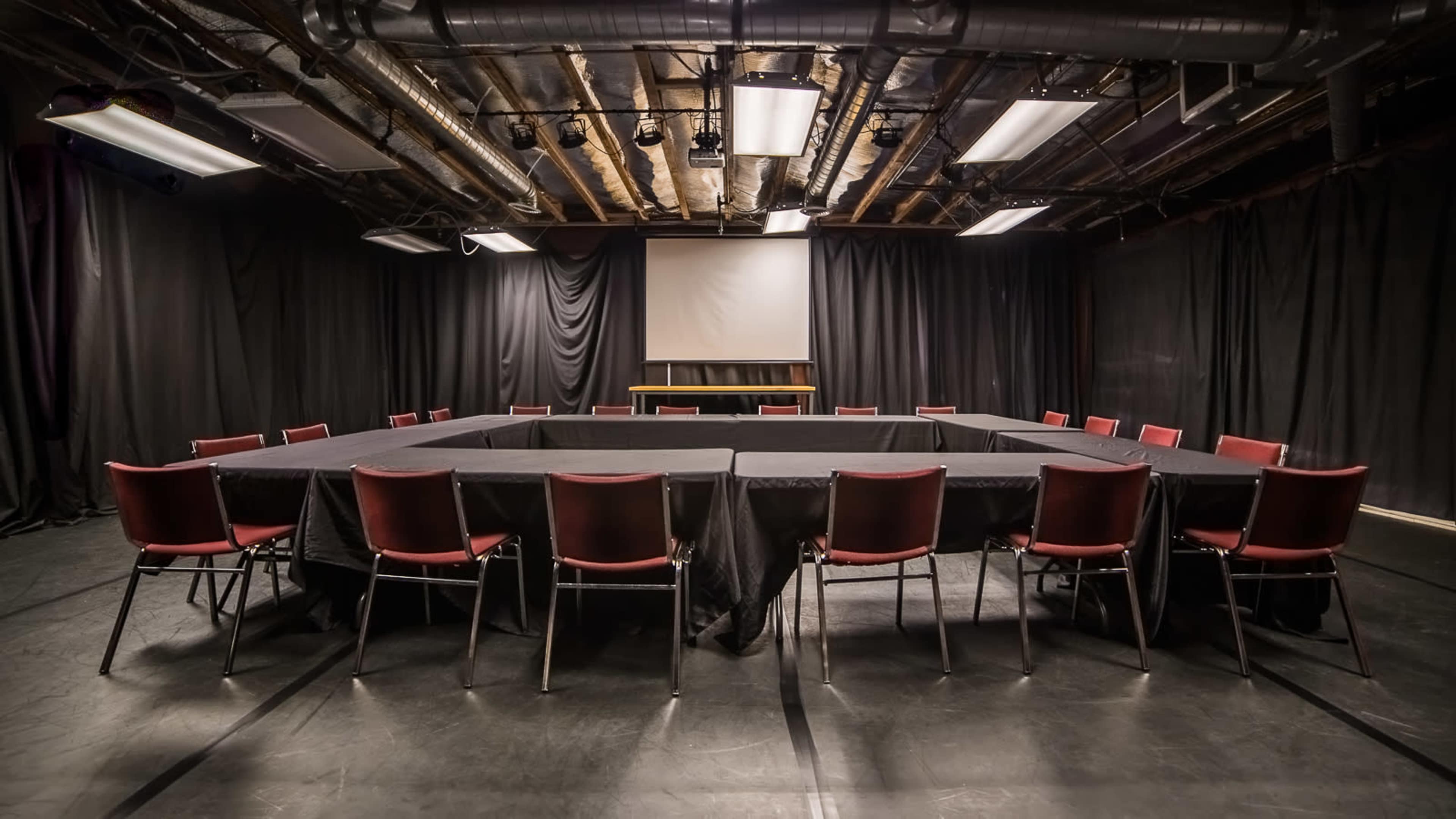 The image shows a conference room with a large rectangular table surrounded by red chairs, set in a space with black curtains and a projector screen at the front.