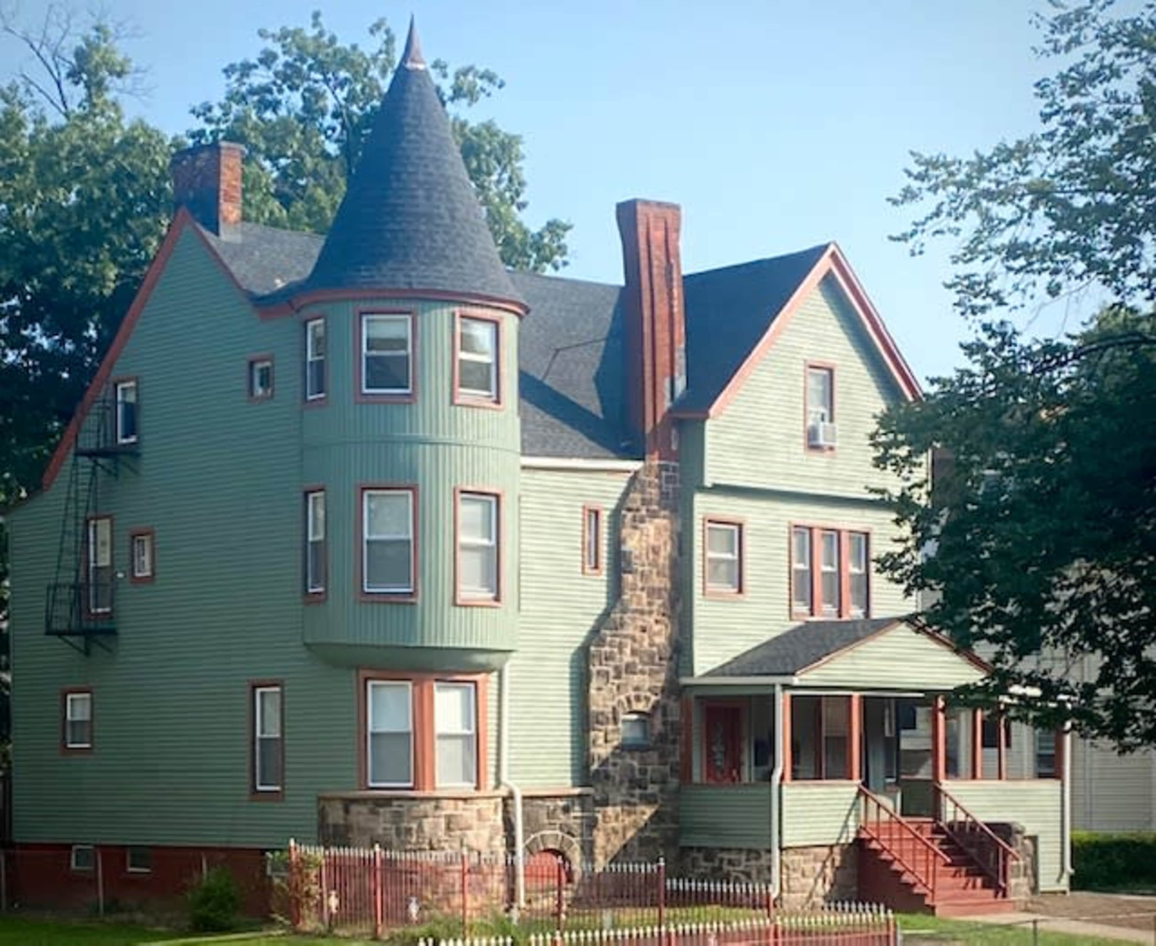 The image shows a large, multi-story house featuring a turret, green siding, and a front porch with steps leading up to it.