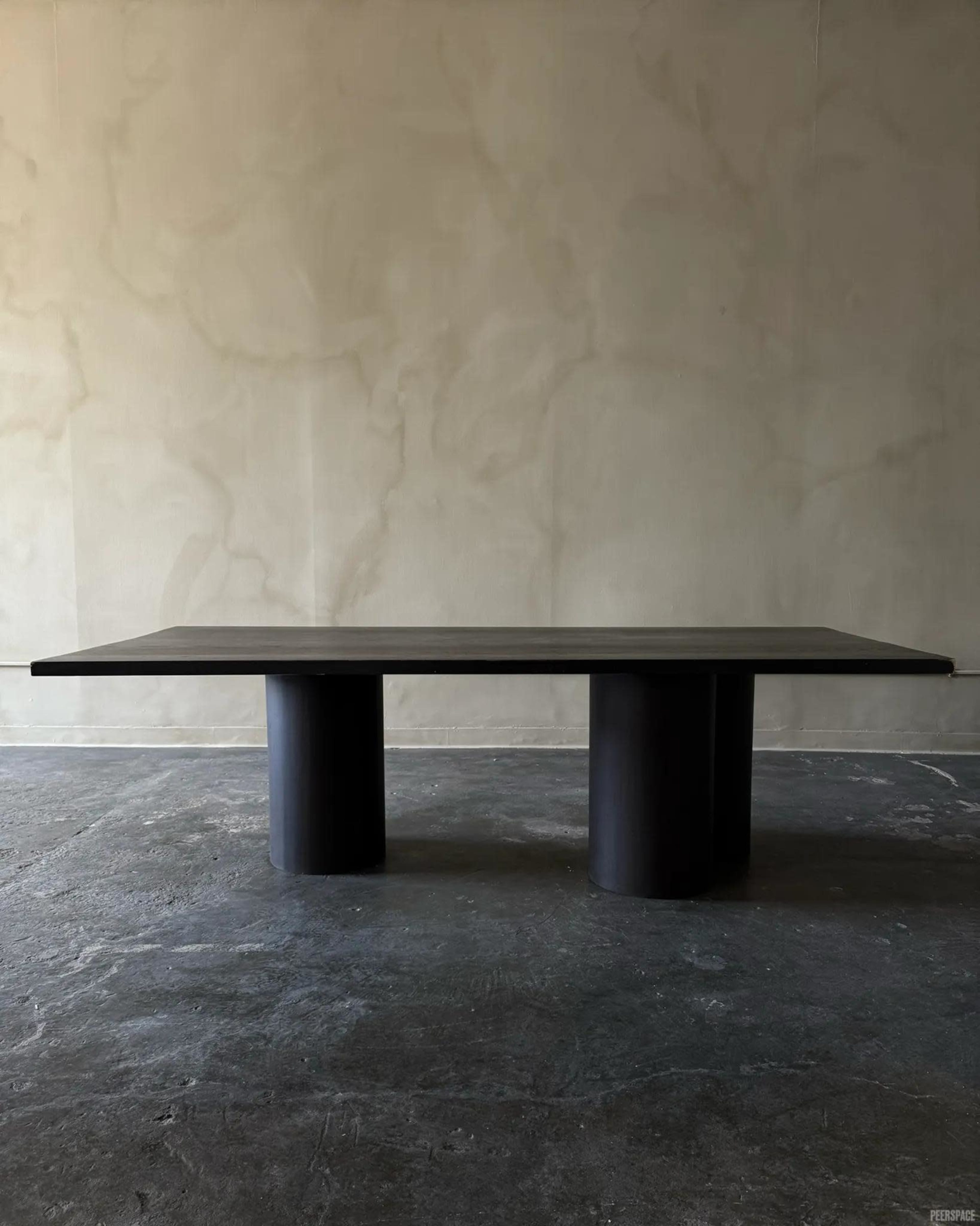 A black minimalist table with a thick top supported by two cylindrical bases stands against a textured gray wall.