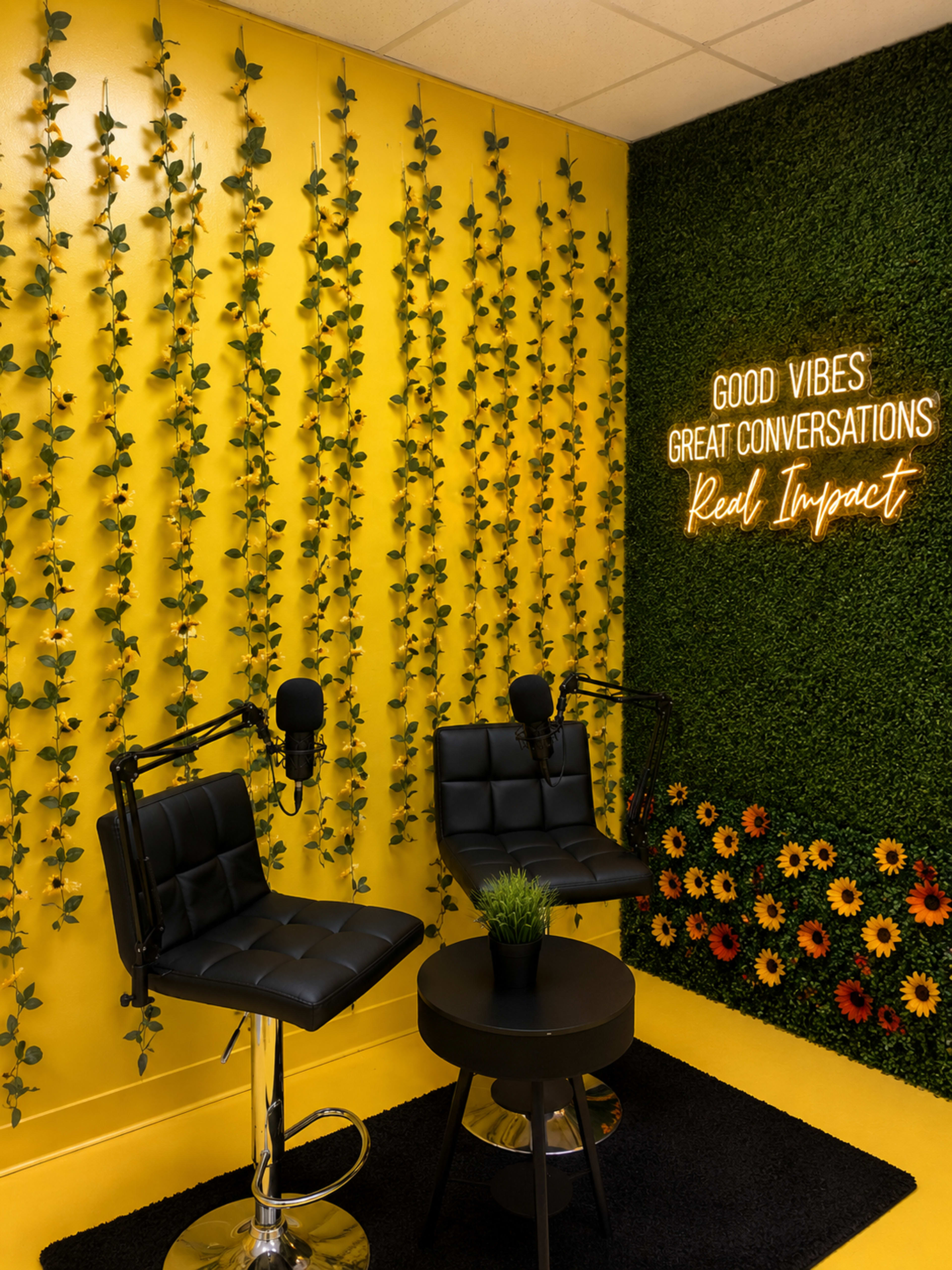 The image shows a vibrant yellow room with two black chairs, a round table, and a wall adorned with artificial greenery and flowers, accompanied by a neon sign that reads "GOOD VIBES GREAT CONVERSATIONS Real Impact."