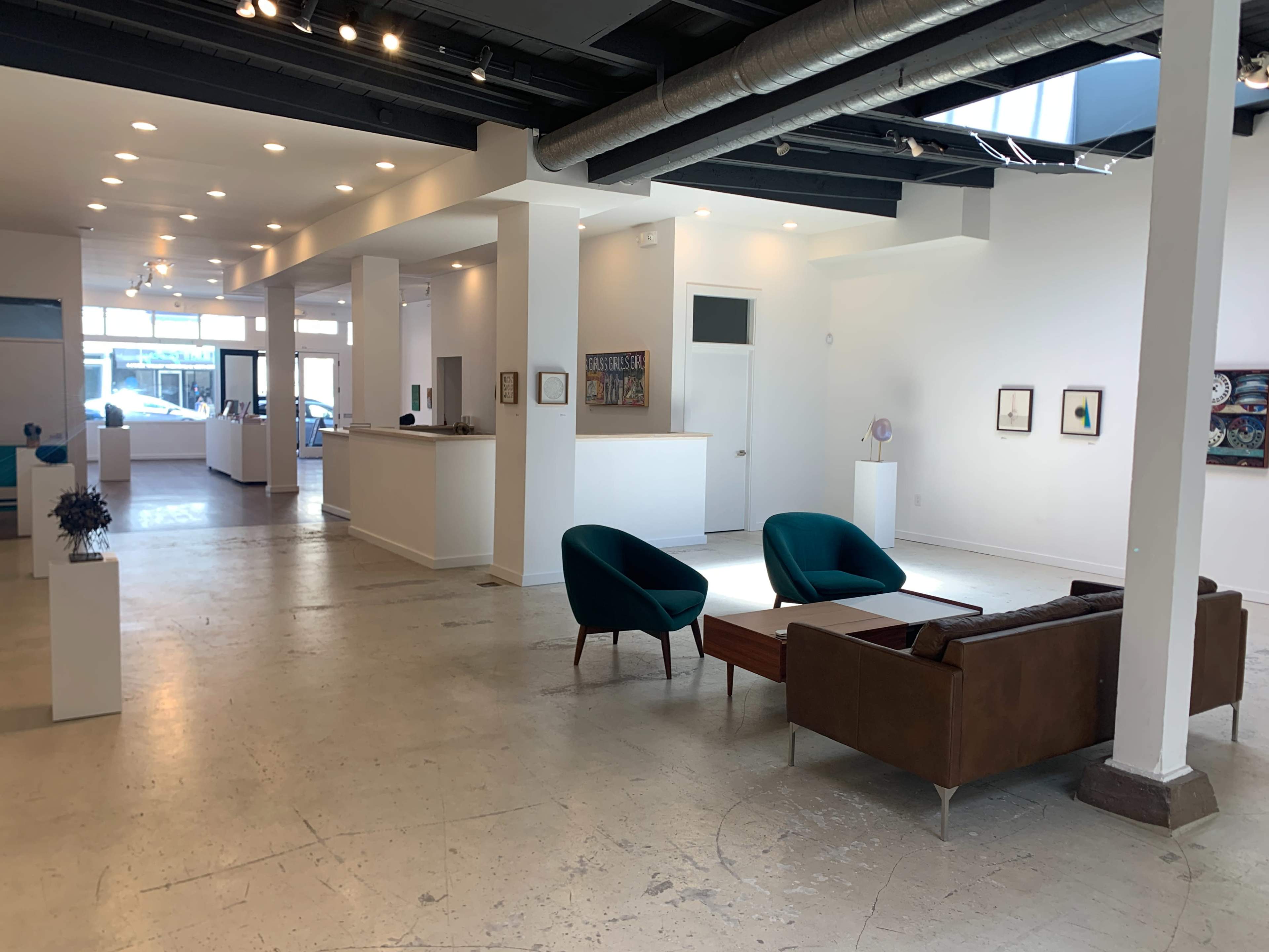 A modern art gallery with white walls, polished concrete floors, and a seating area featuring two teal chairs and a brown couch.