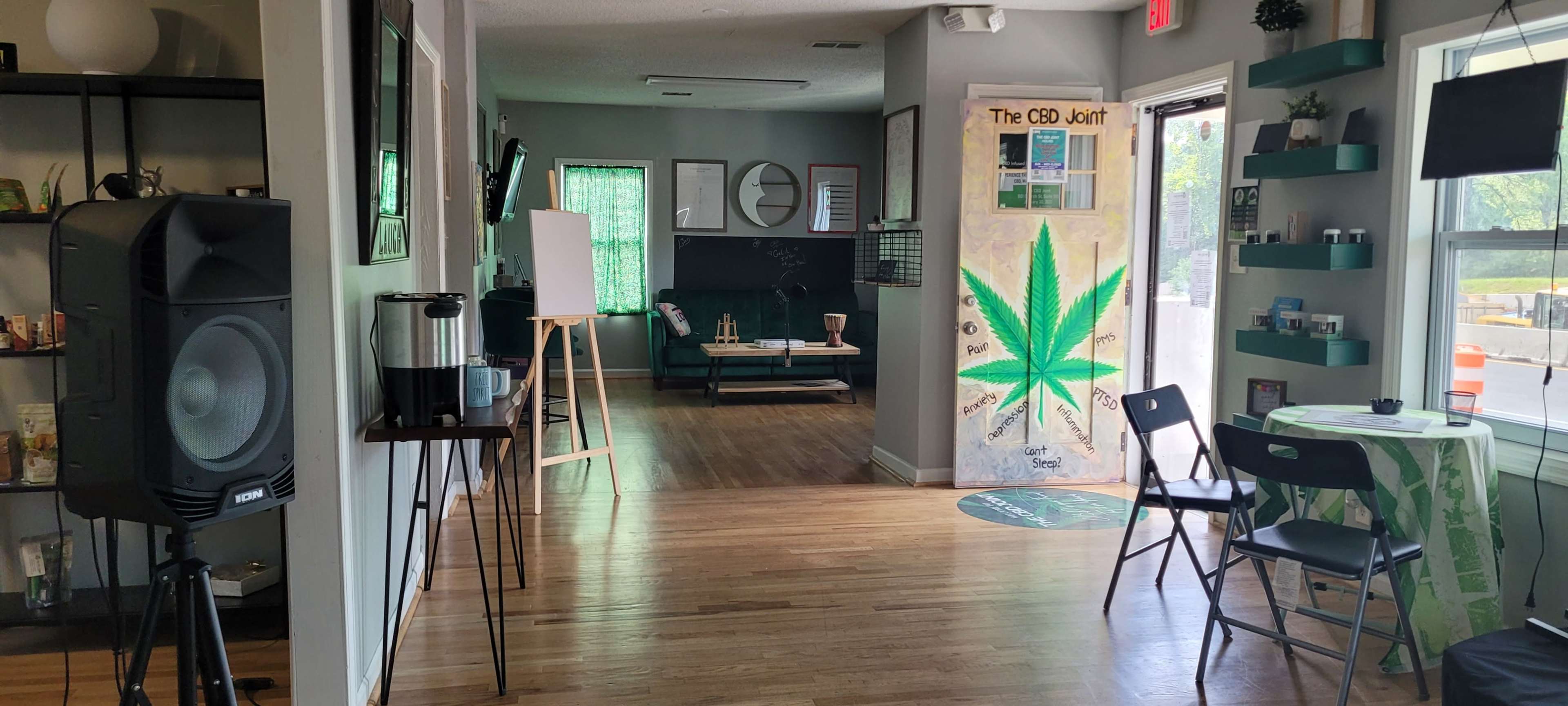 The interior of a cannabis-themed shop features a waiting area with chairs, a large green cannabis leaf mural, and various decorations along the walls.