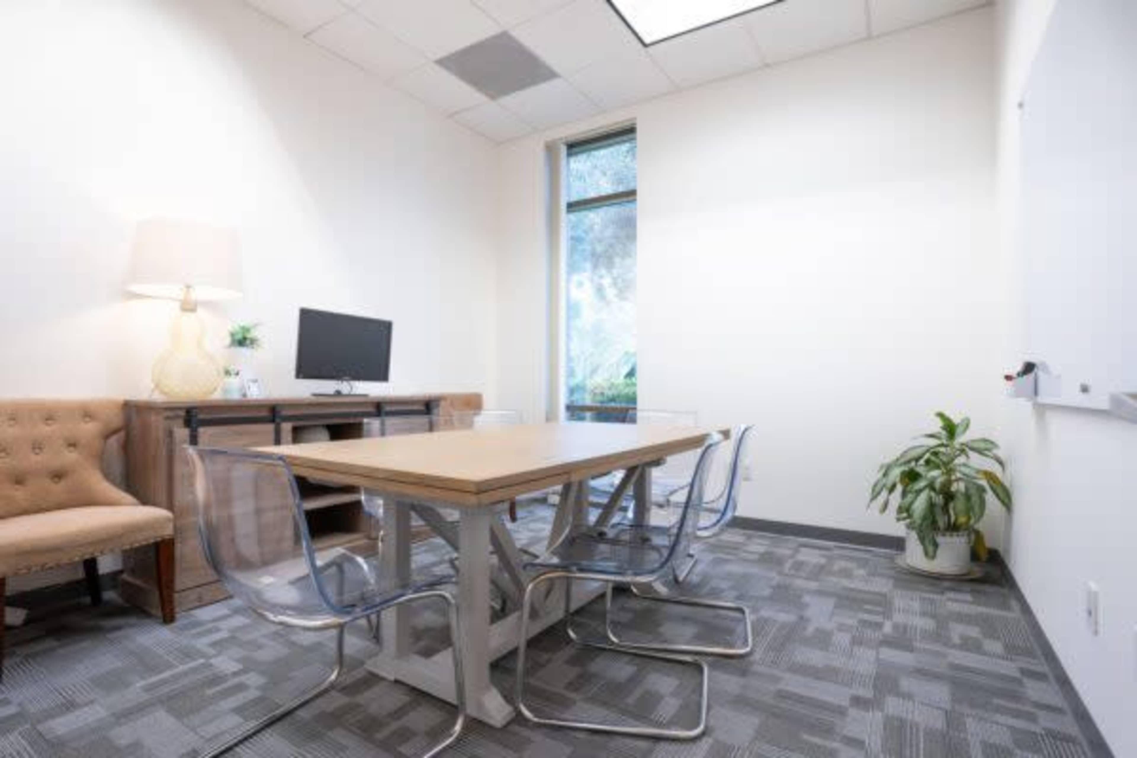 The image shows a small meeting room with a wooden table surrounded by clear chairs, a wall-mounted monitor, a lamp, a plant, and a large window.