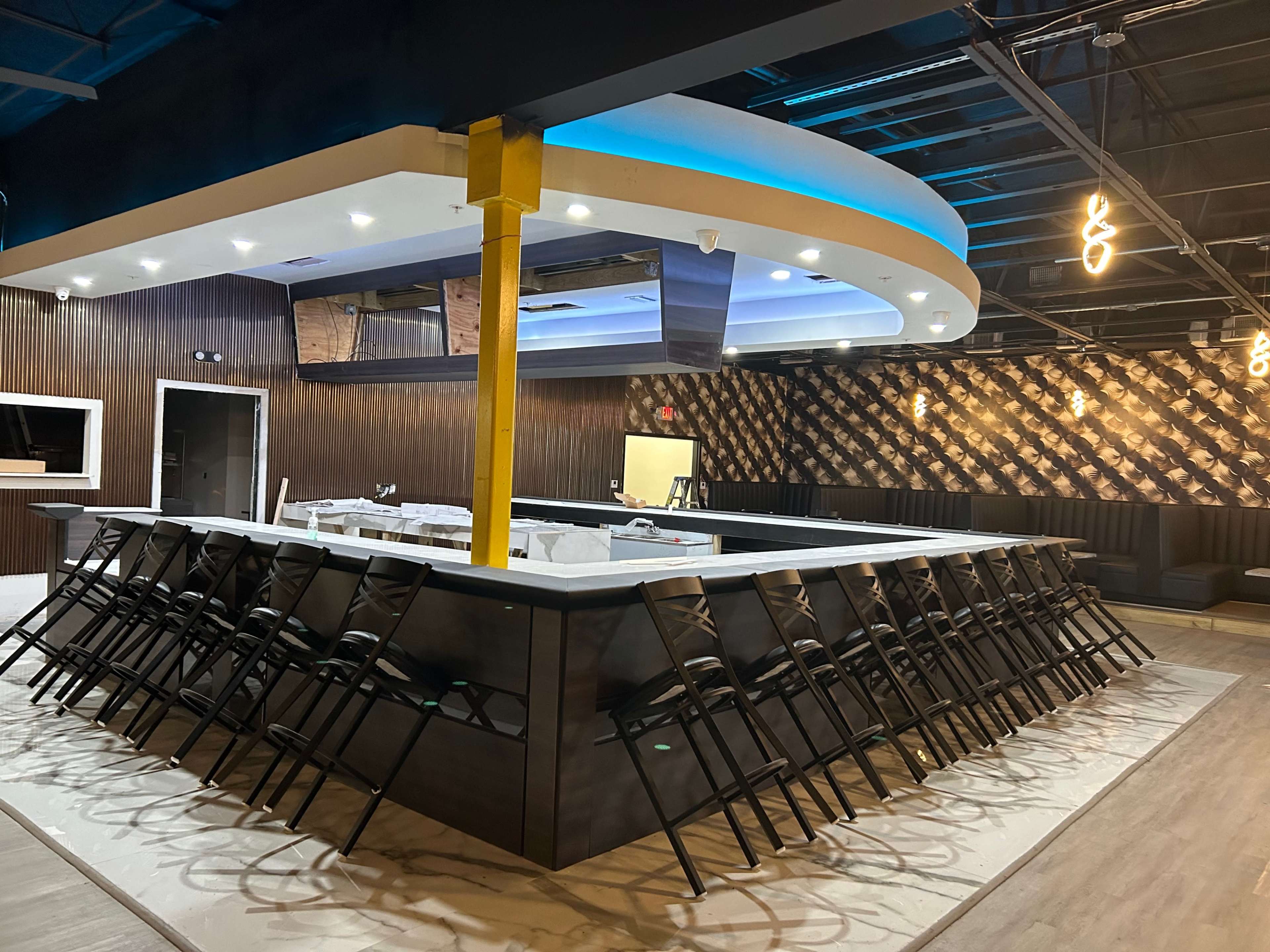 A large, modern bar area features a sweeping counter with multiple stools arranged neatly, set against a backdrop of dark wood and stylish lighting.