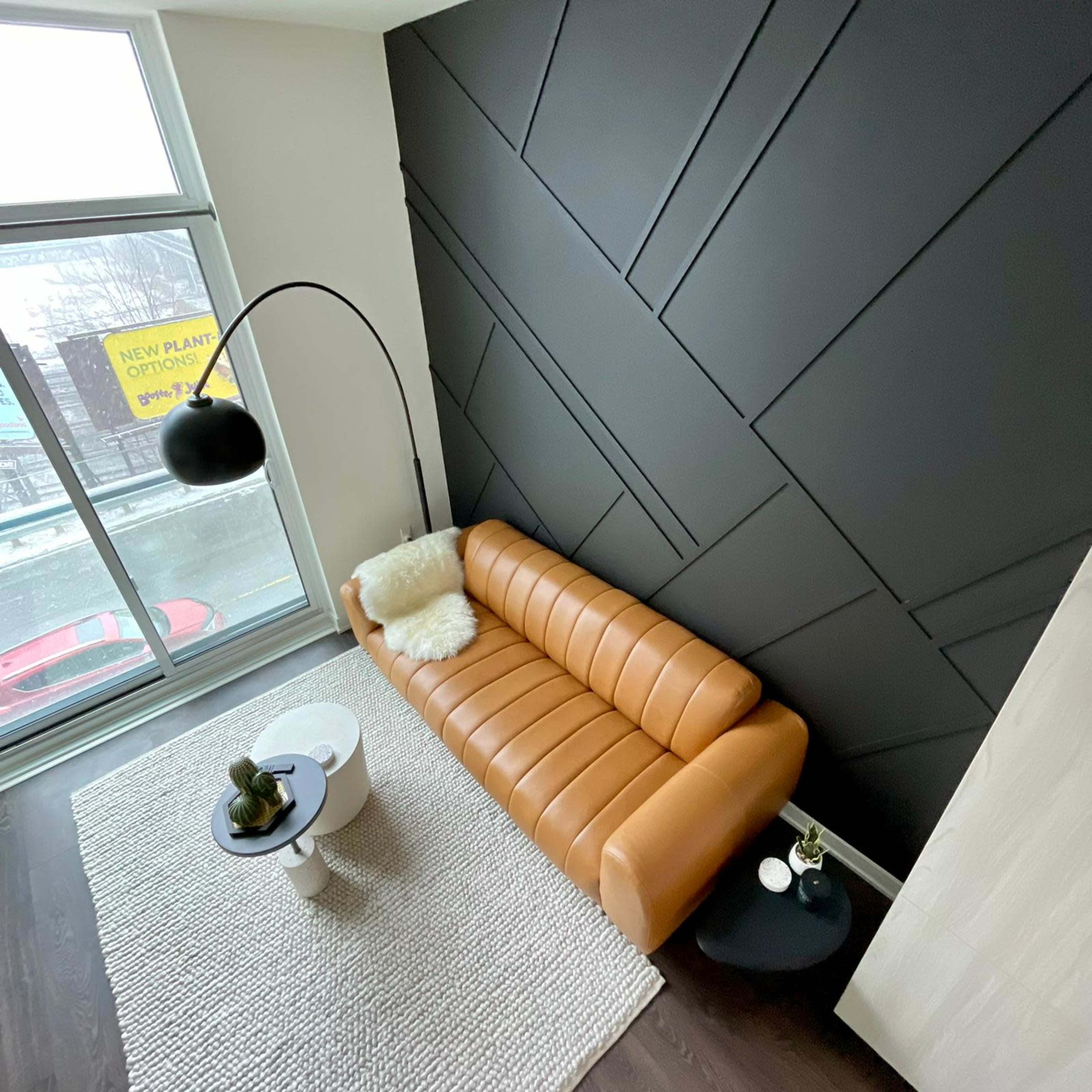 A modern living room with a brown leather sofa, a round white coffee table, a small black side table, and a floor lamp against a geometric black accent wall.