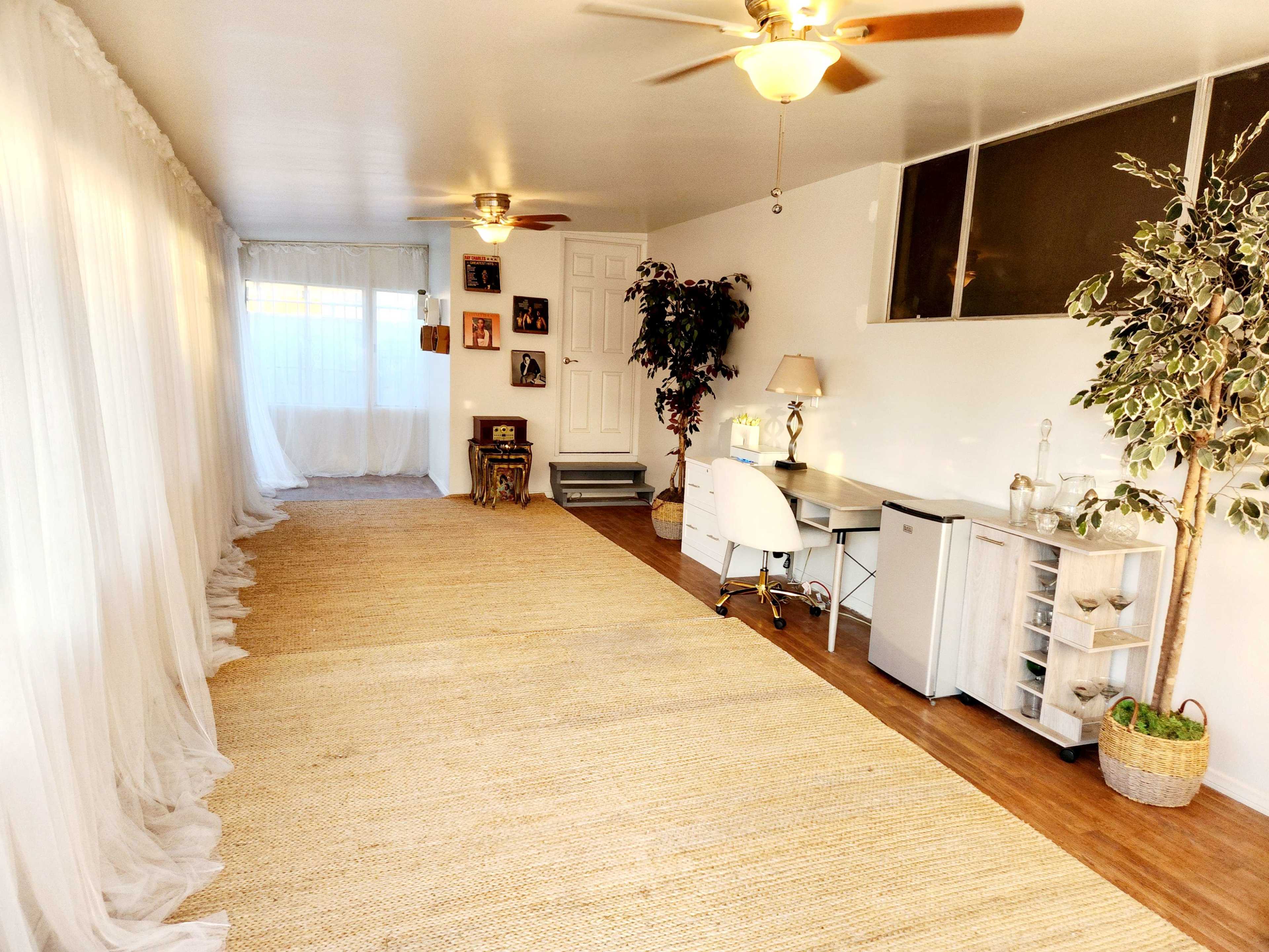 A spacious room with natural light, featuring a jute rug, a desk with a chair, and decorative plants.