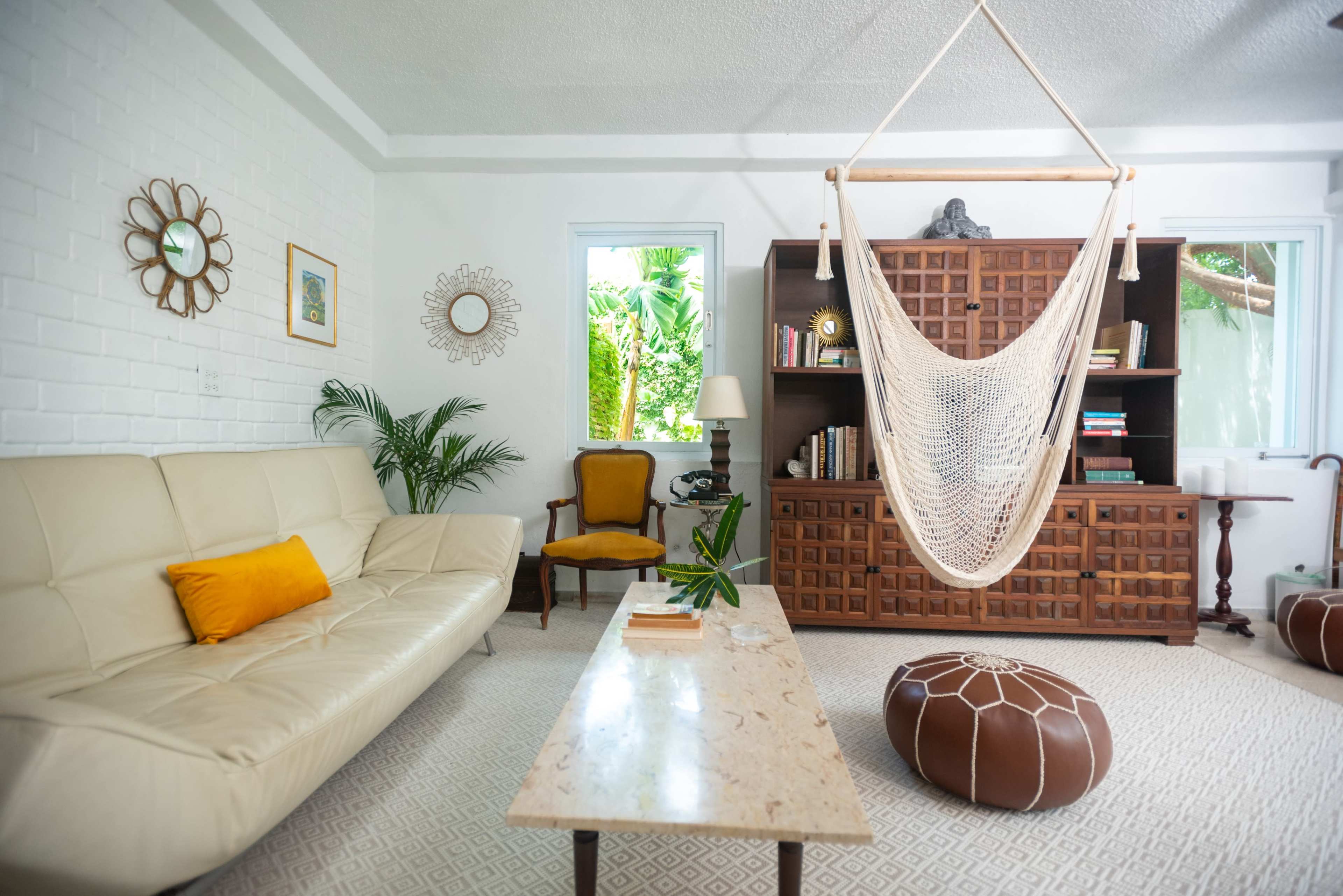 The image shows a bright living room with a cream-colored sofa, a wooden bookshelf, a hammock hanging from the ceiling, and a coffee table in front.
