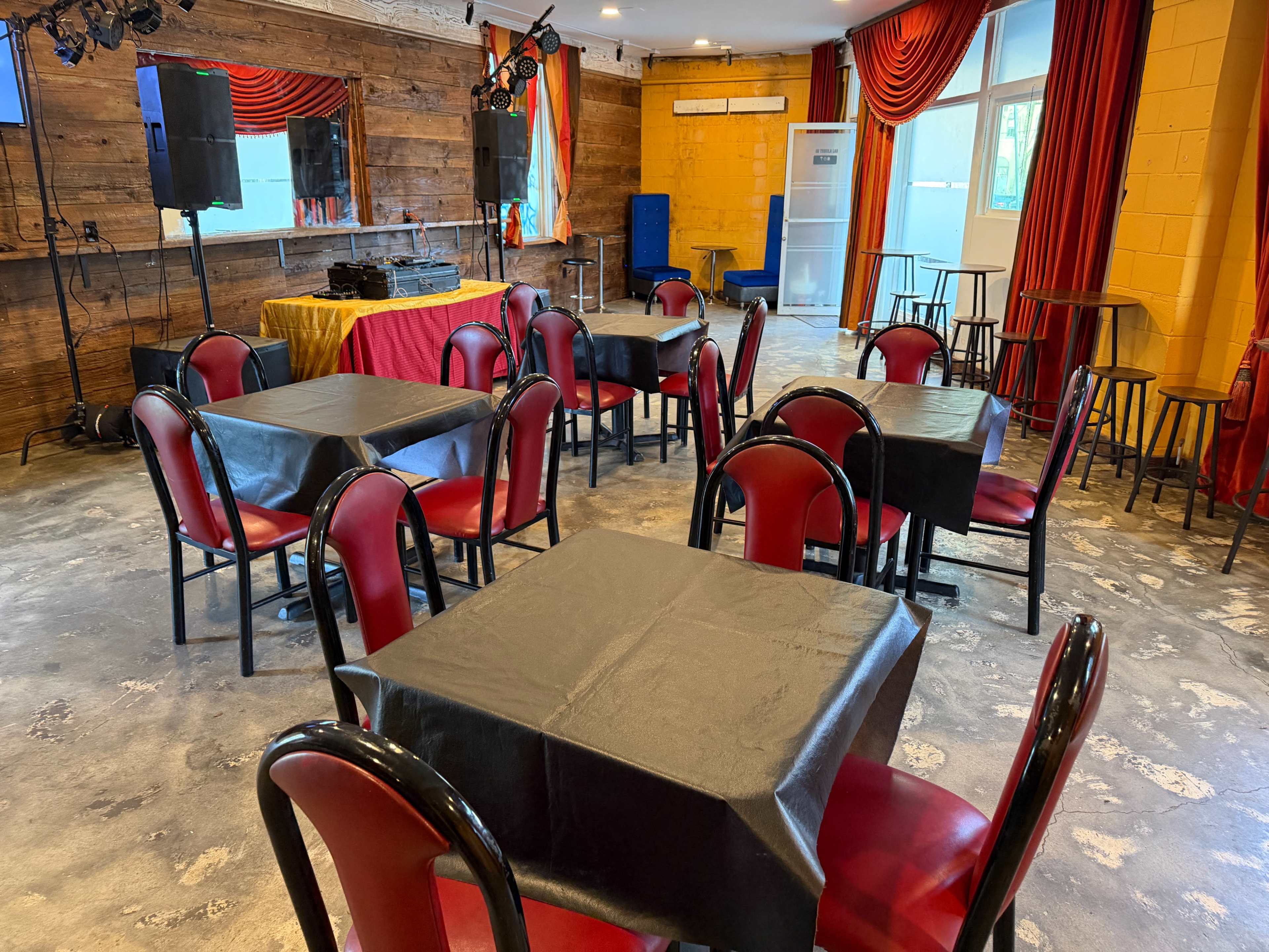 The image shows an empty event space with several black tables and red chairs arranged, along with audio equipment and stage lights in the background.