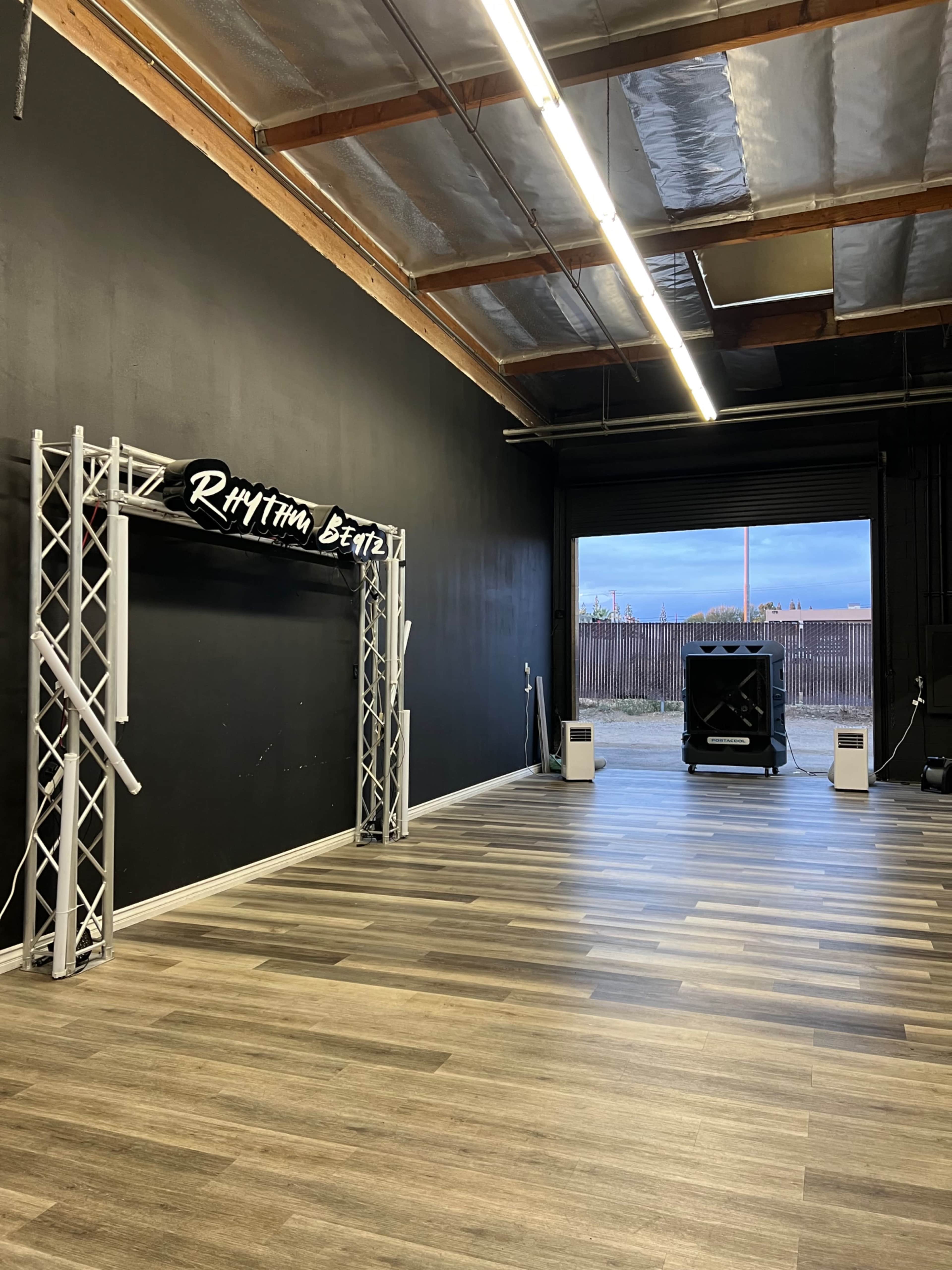 The image shows a large, empty studio space with wooden flooring and a black wall featuring a decorative archway labeled "RHYTHM BEATS."