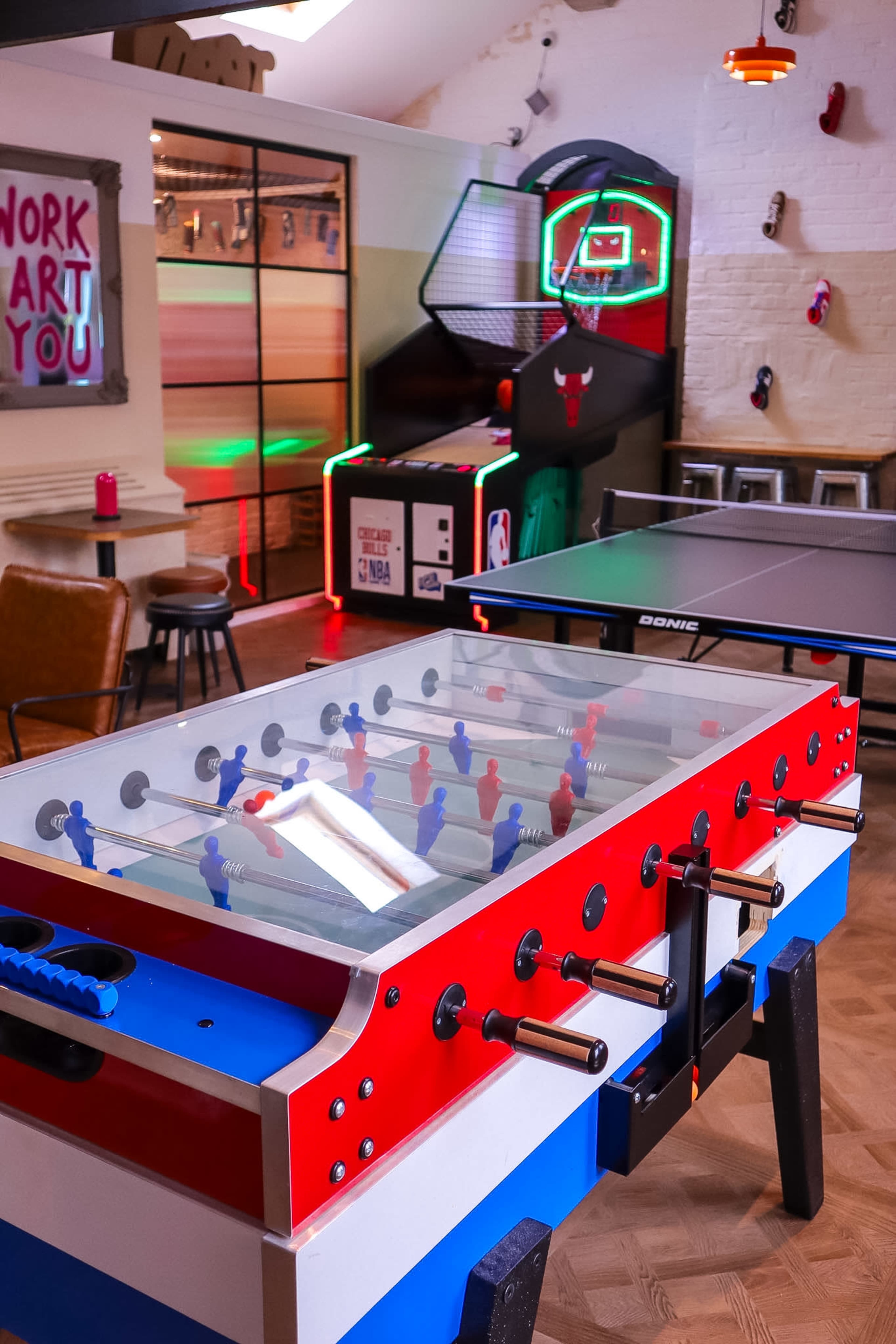 The image shows a game room featuring a foosball table, a ping pong table, and a basketball arcade game, with colorful lighting and decor.