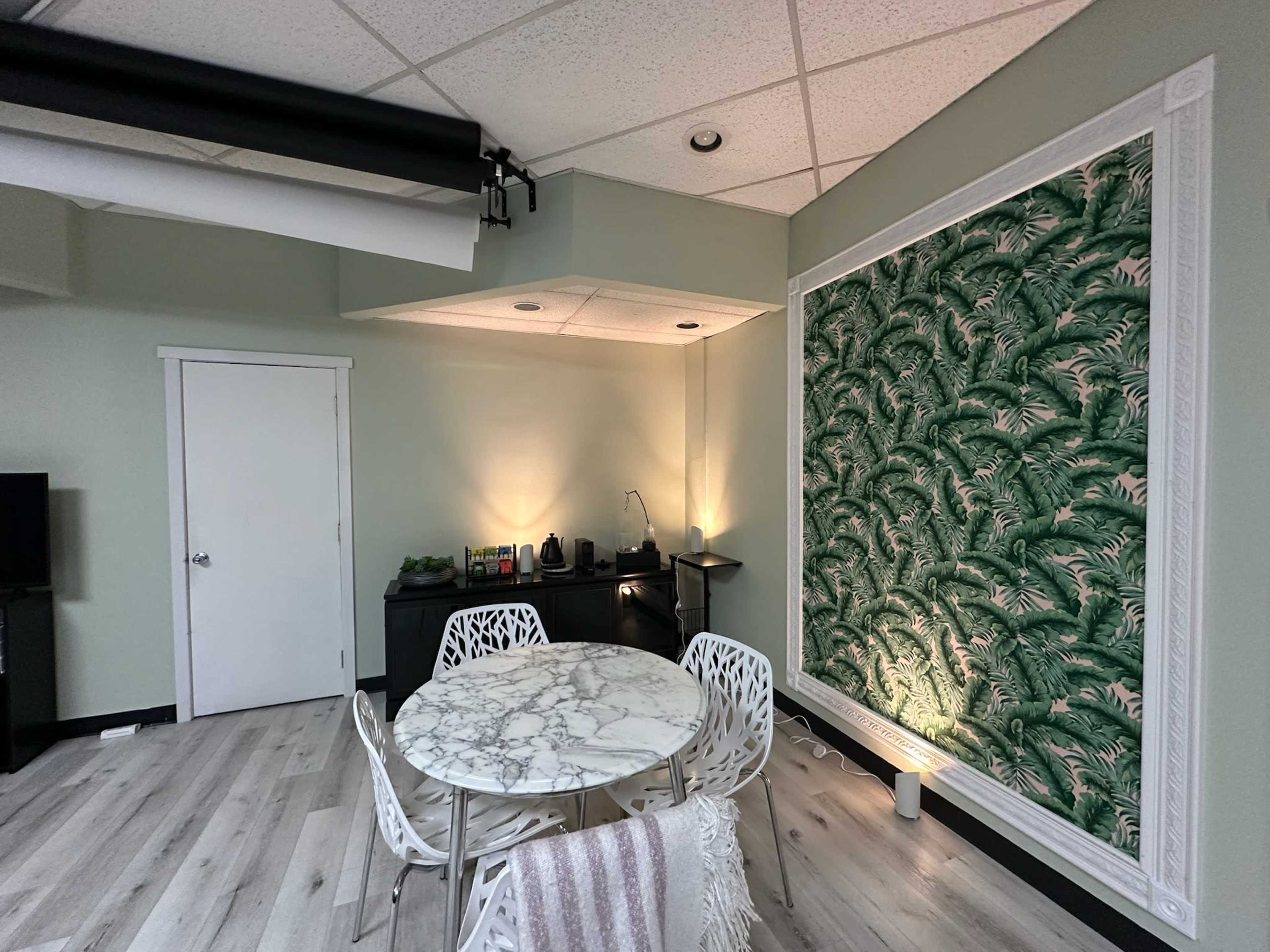 The image shows a modern room featuring a marble-top table with white chairs, a black cabinet, and a lush green palm leaf wallpaper on one wall.