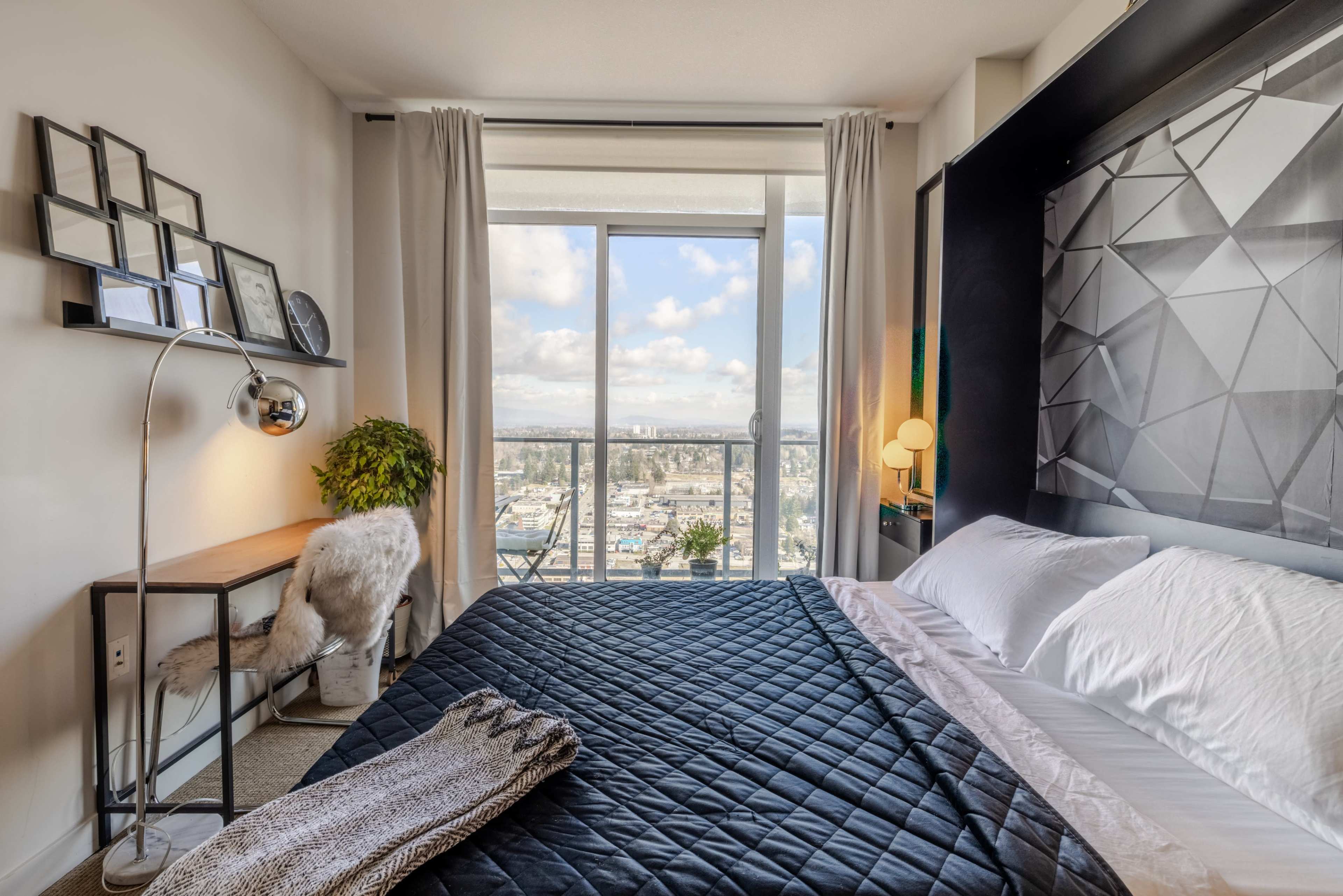 A modern bedroom features a large window with city views, a bed with a quilted black cover, and a desk with a lamp and decorative elements.