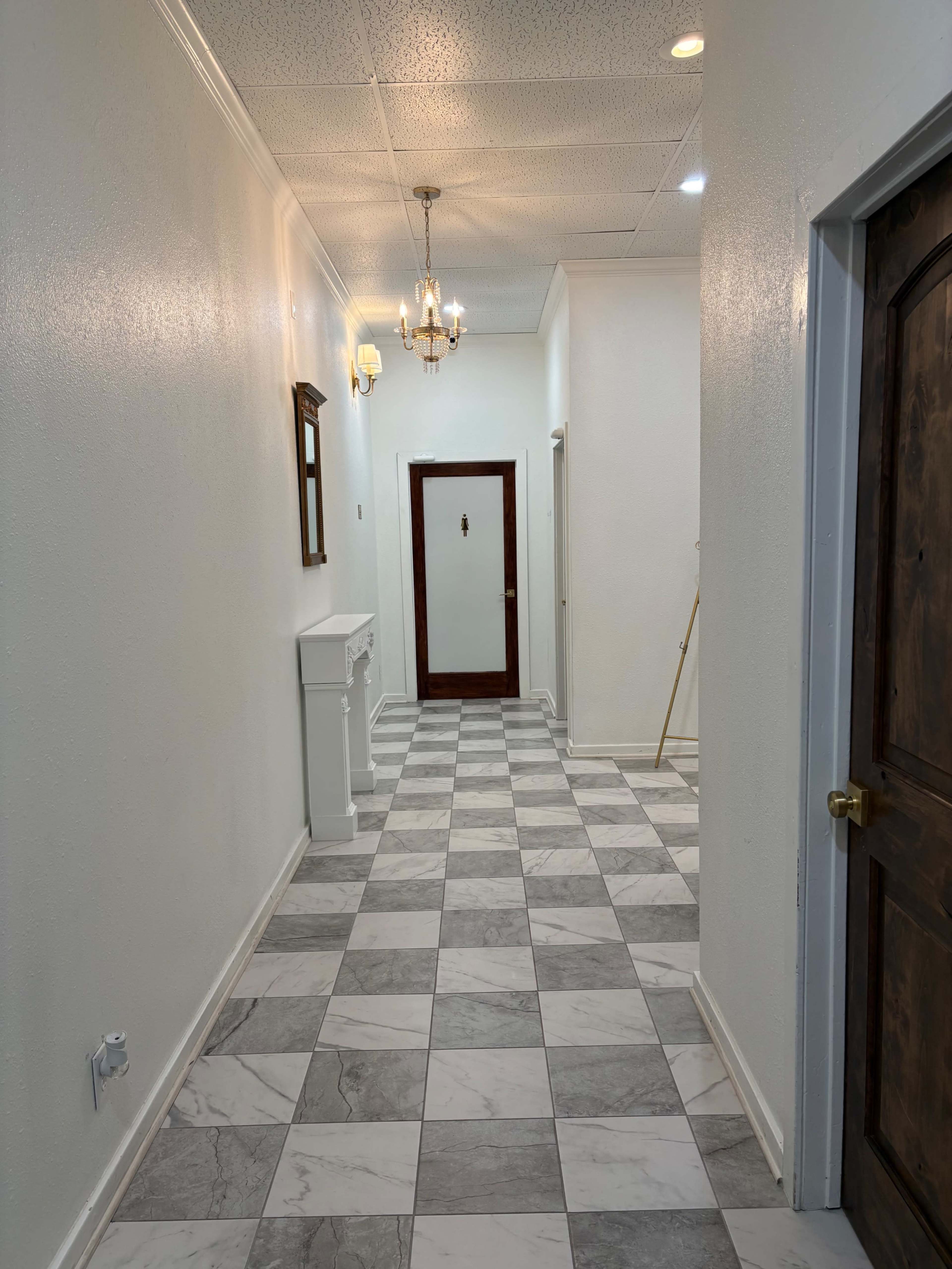 The image shows a spacious, well-lit hallway with a checkered marble floor, white walls, and a wooden door at the end.