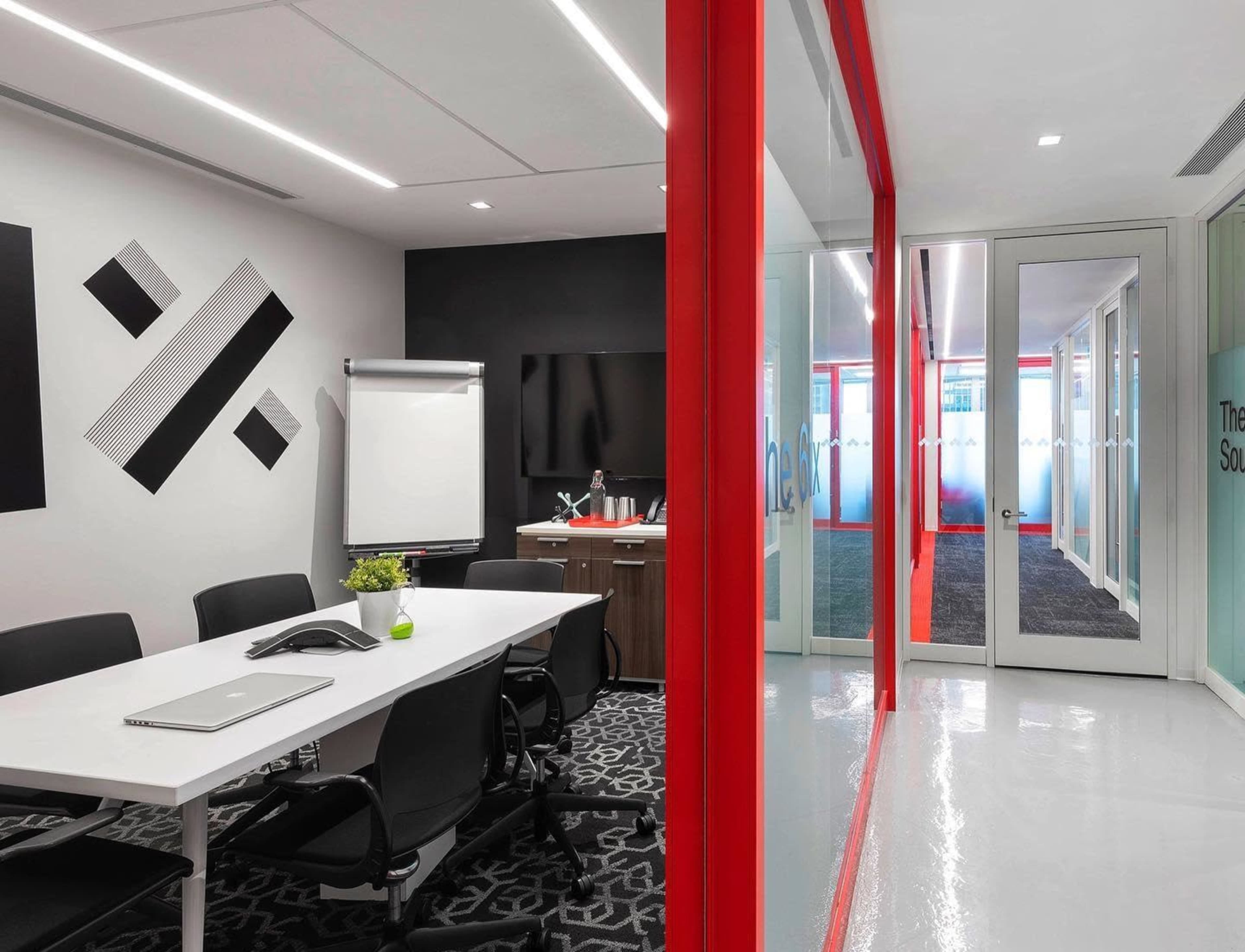 A modern meeting room with a white table and black chairs, featuring a large screen and a glass wall leading to a hallway with red accents.