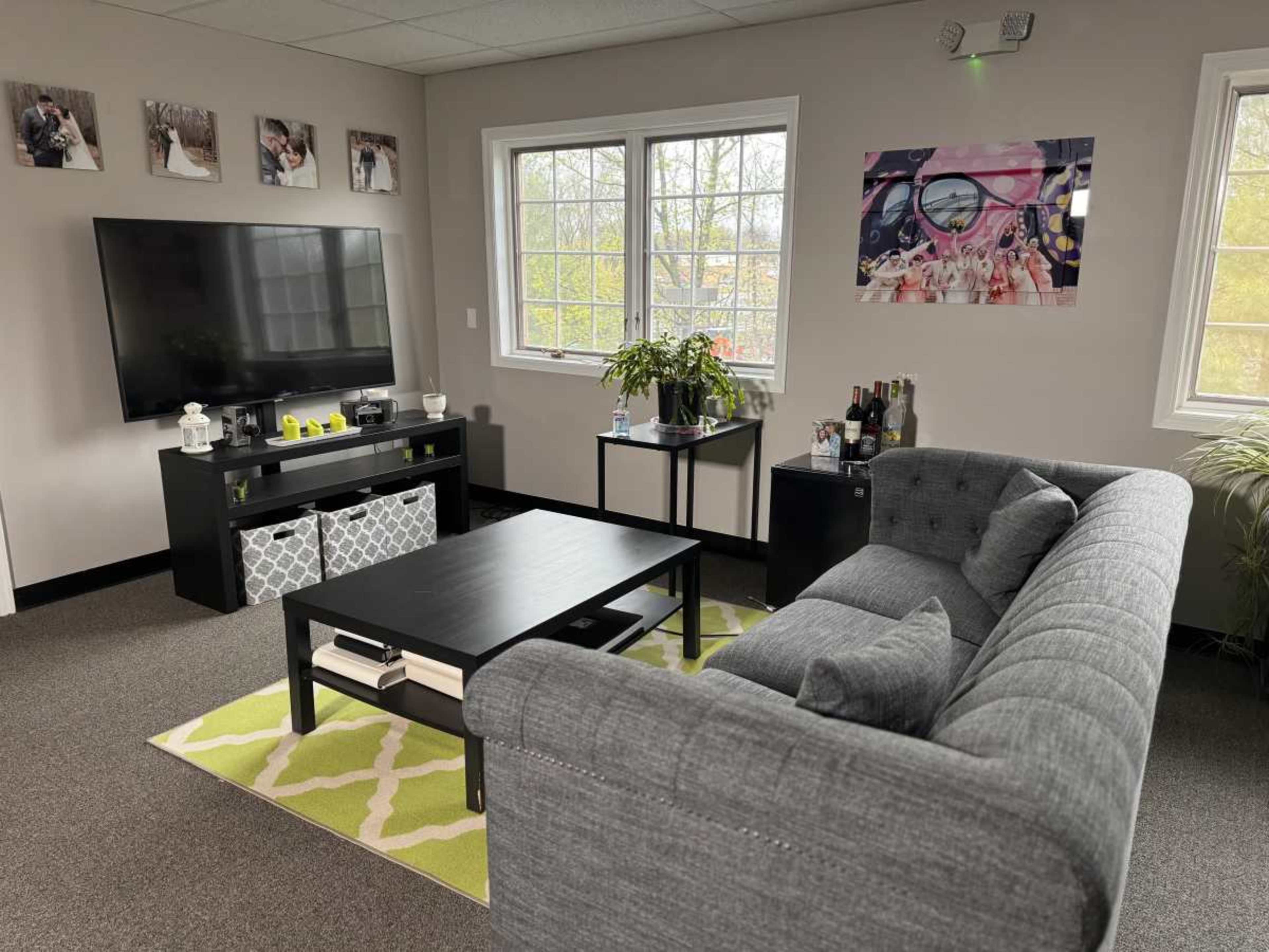 The image shows a modern living room with a gray couch, a black coffee table, a television mounted on the wall, and large windows letting in natural light.