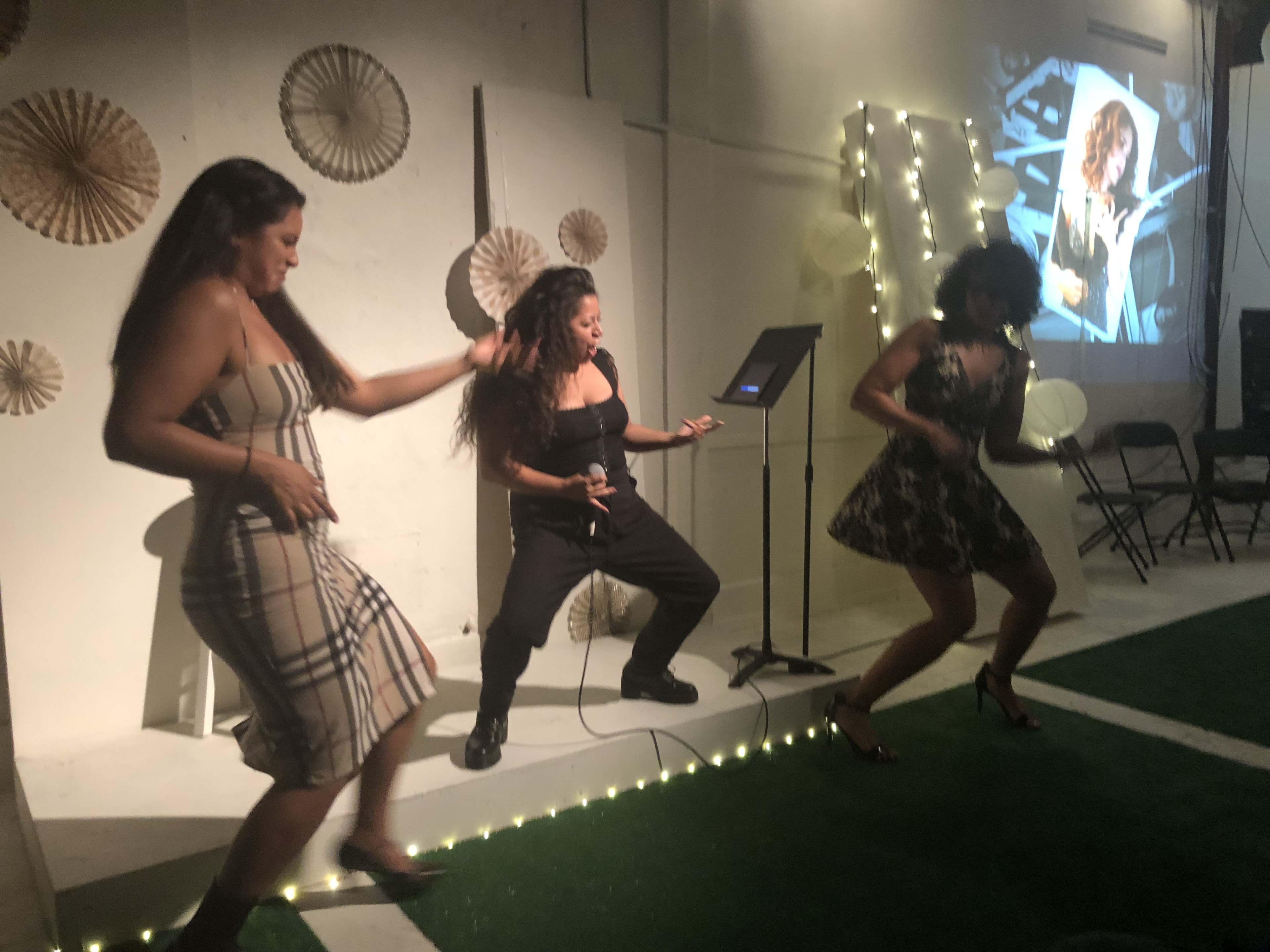 Three women perform a dance routine on stage with a microphone and a backdrop featuring a projected image.