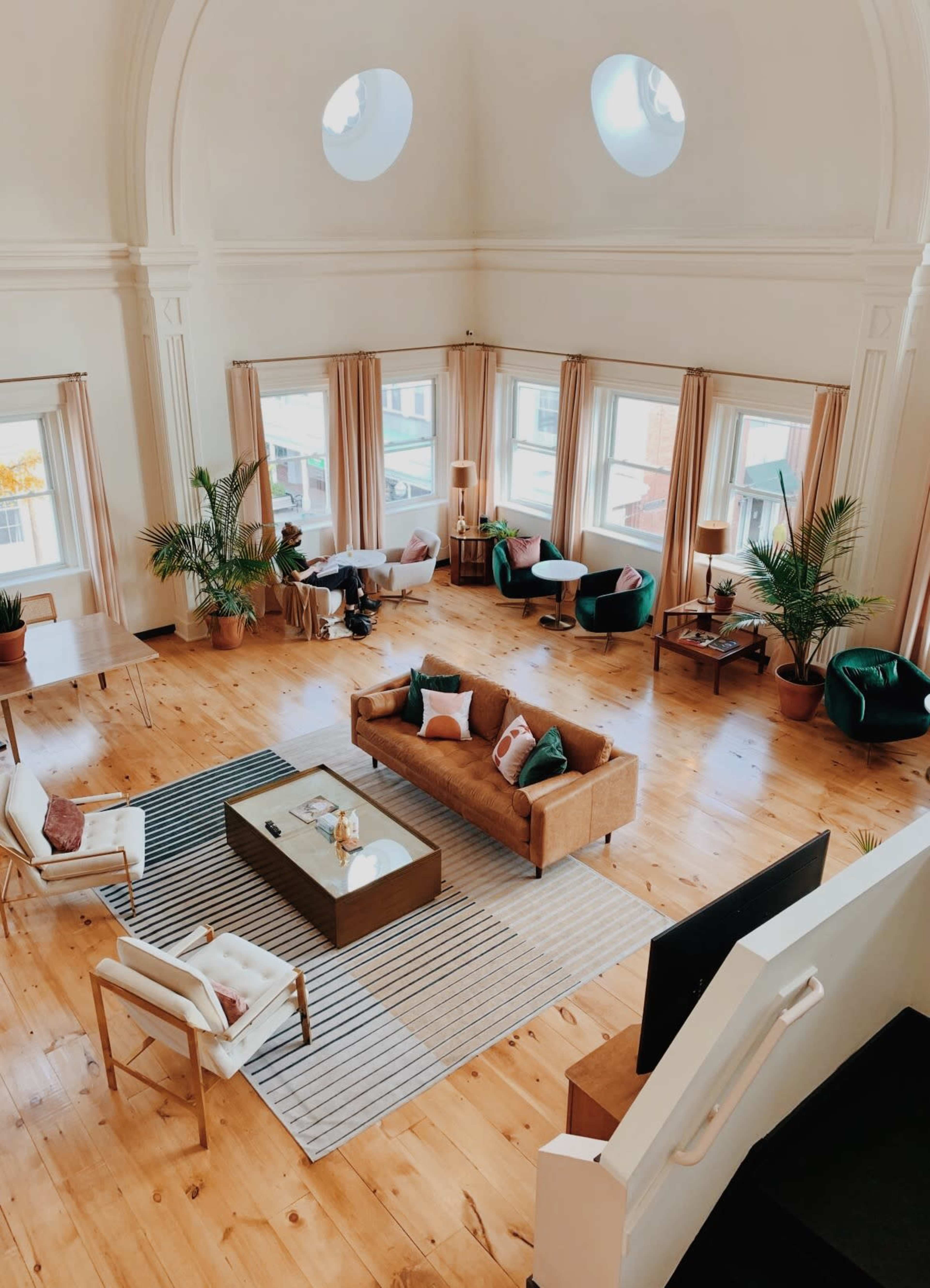 A spacious, elegantly designed living area with large windows, wooden flooring, and a combination of seating arrangements surrounded by plants.