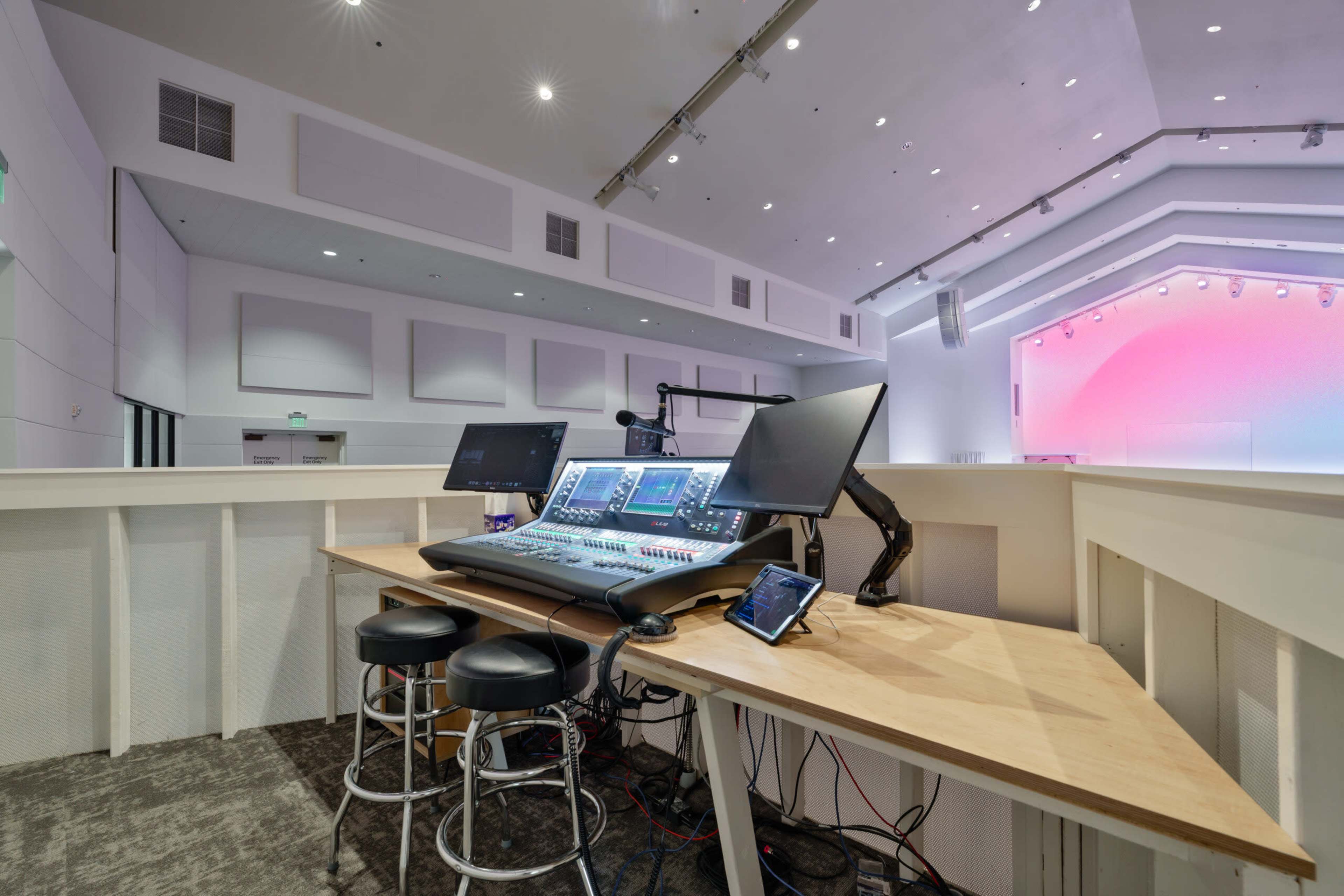 The image shows a modern audio control station with multiple screens and a sound mixing console, positioned in a spacious venue.
