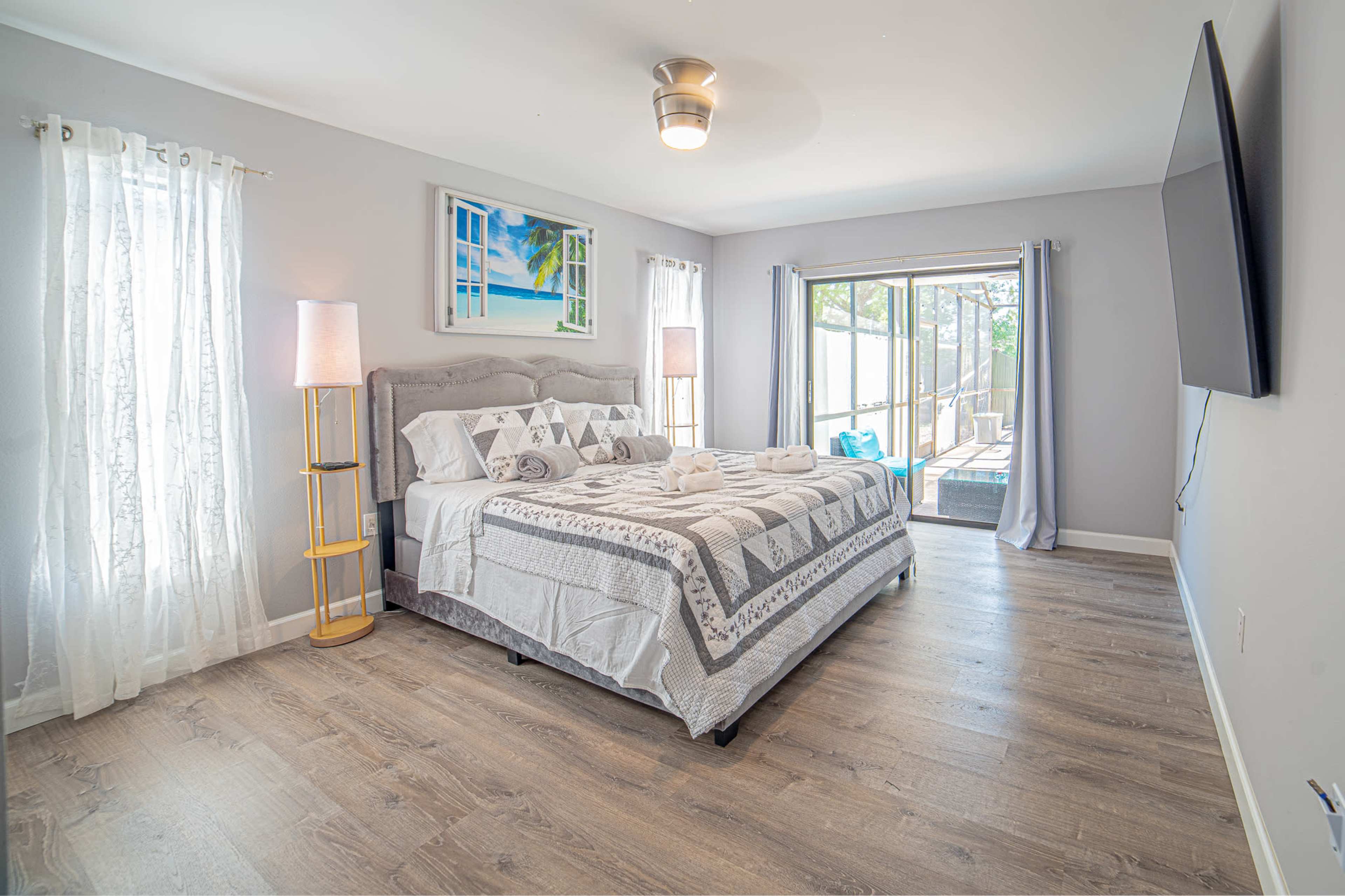 The image shows a neatly arranged bedroom featuring a large bed with a patterned quilt, two lamps, and a sliding glass door leading to an outdoor area.