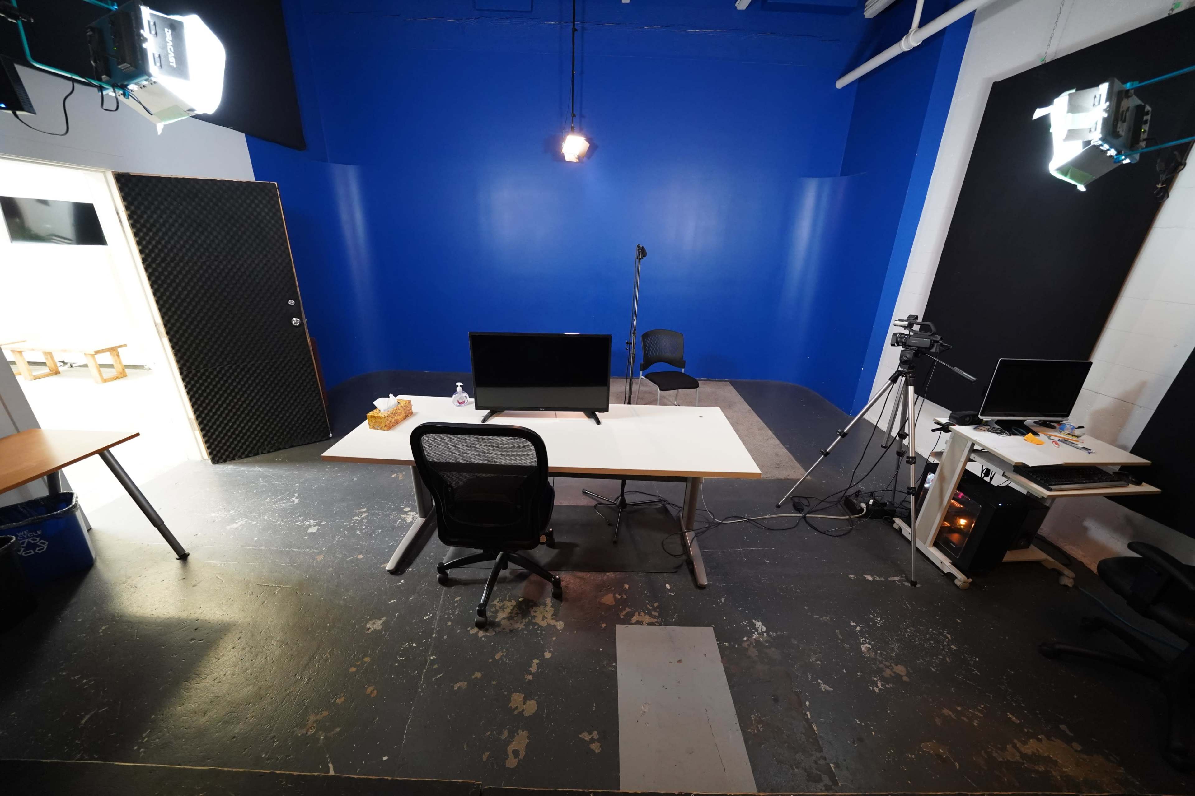 The image shows a sparsely furnished studio with a blue wall, a desk with a monitor, a chair, and lighting equipment set up for recording.