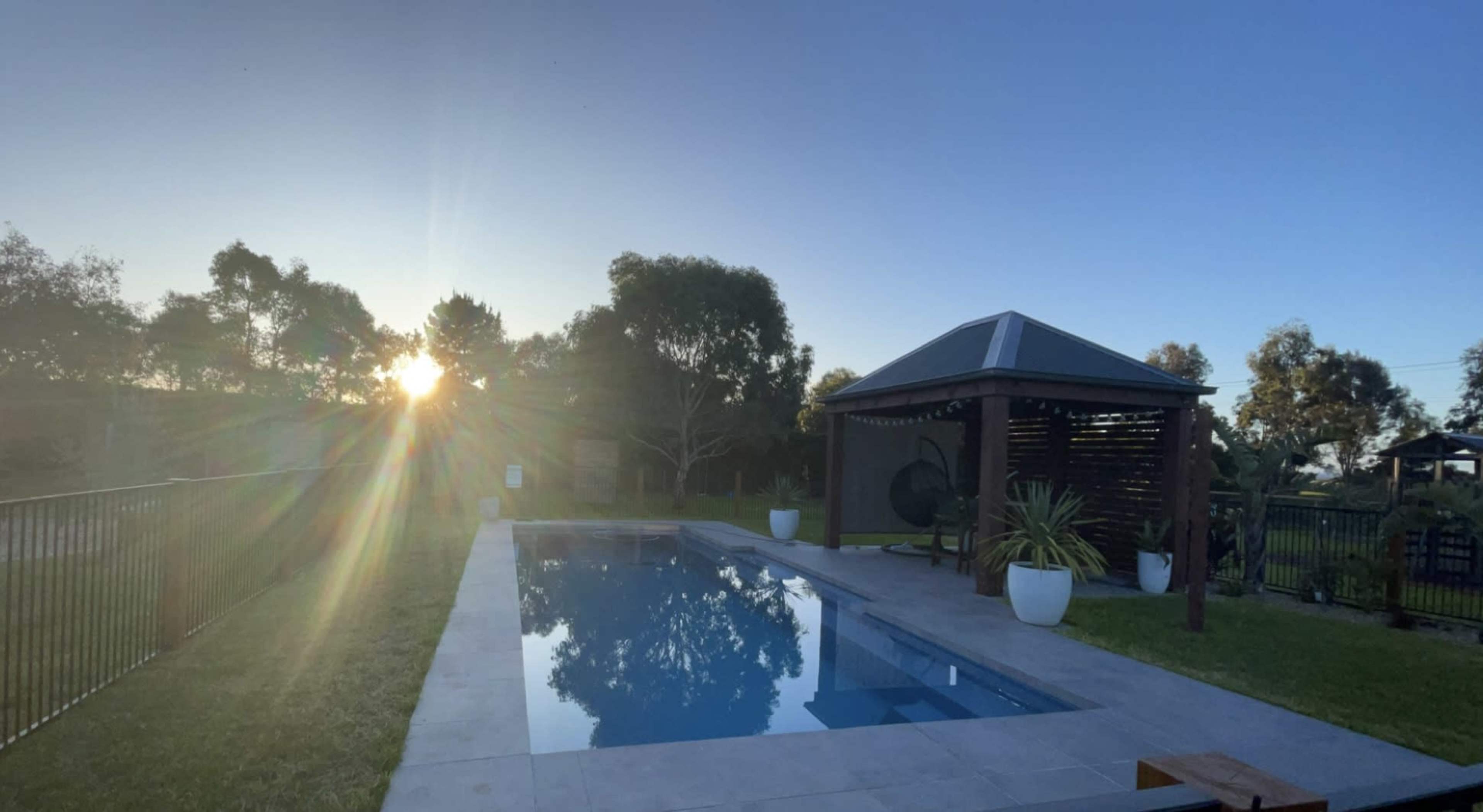 A rectangular swimming pool beside a wooden gazebo, with the sun setting in the background.