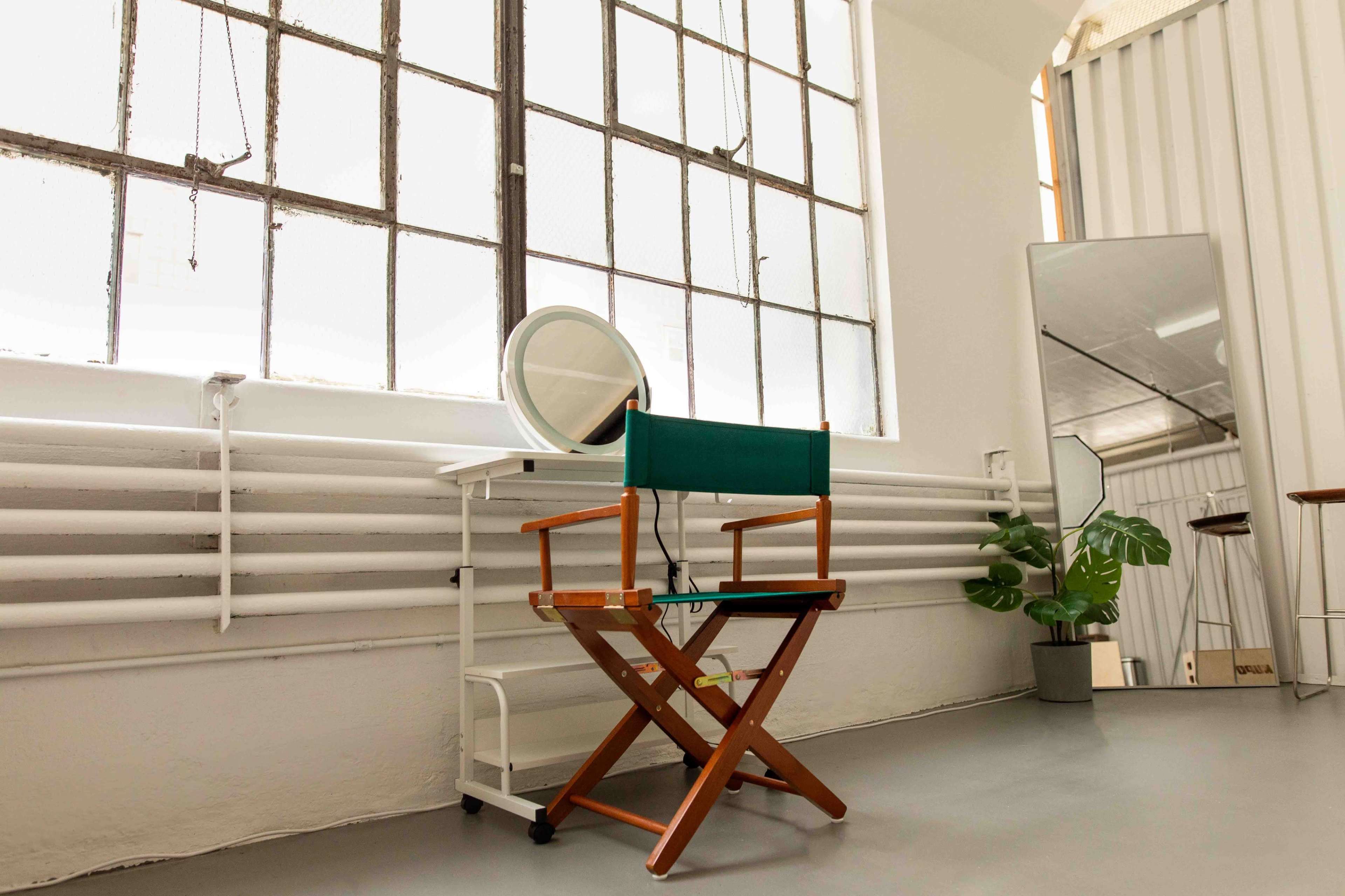 A minimalist interior space with a wooden director's chair, a round mirror on a small cart, a tall mirror, and a potted plant near large windows.
