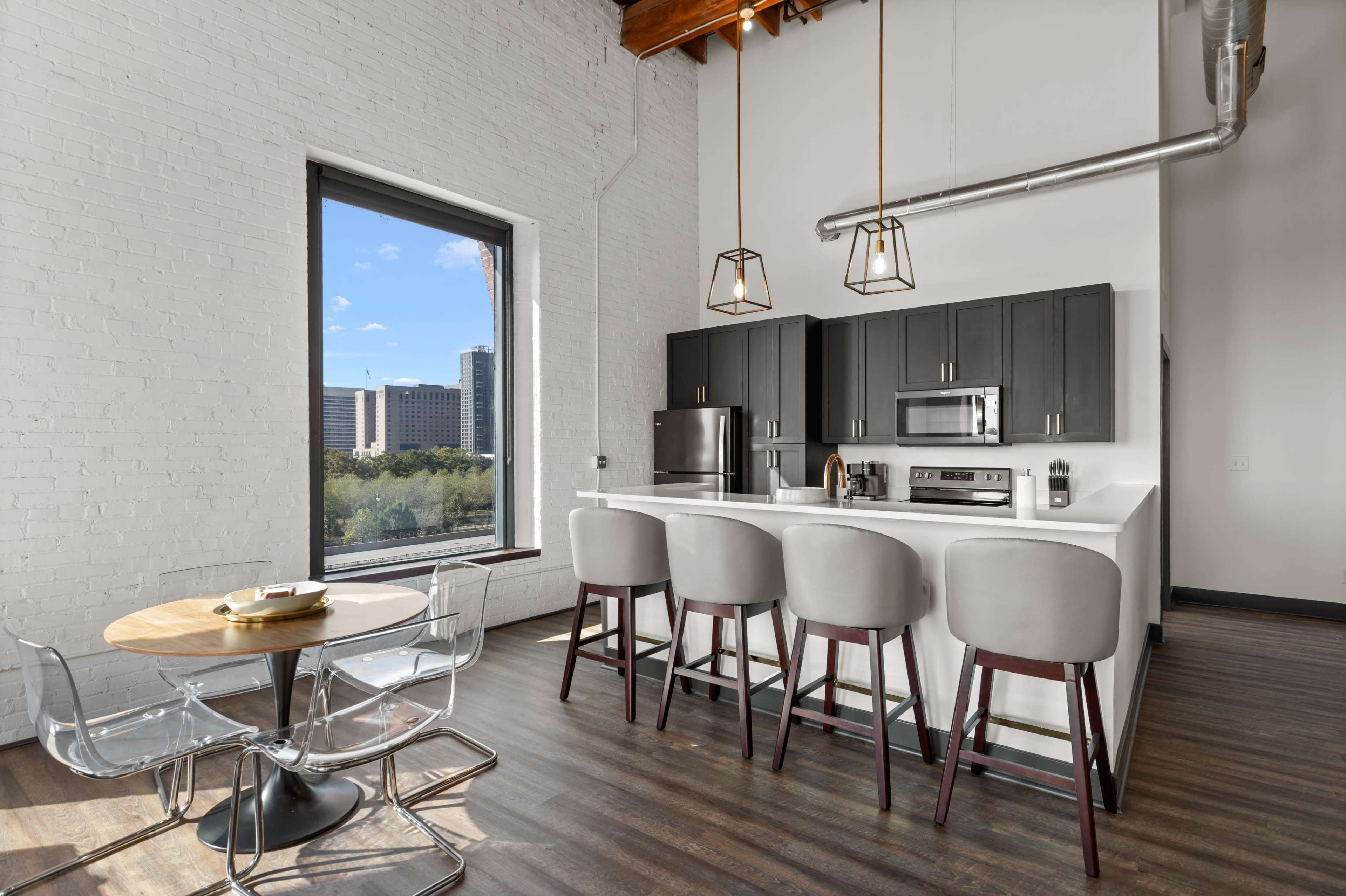 The image shows a modern kitchen with a breakfast bar and a dining area featuring a round table and transparent chairs, set against a backdrop of large windows revealing an urban view.