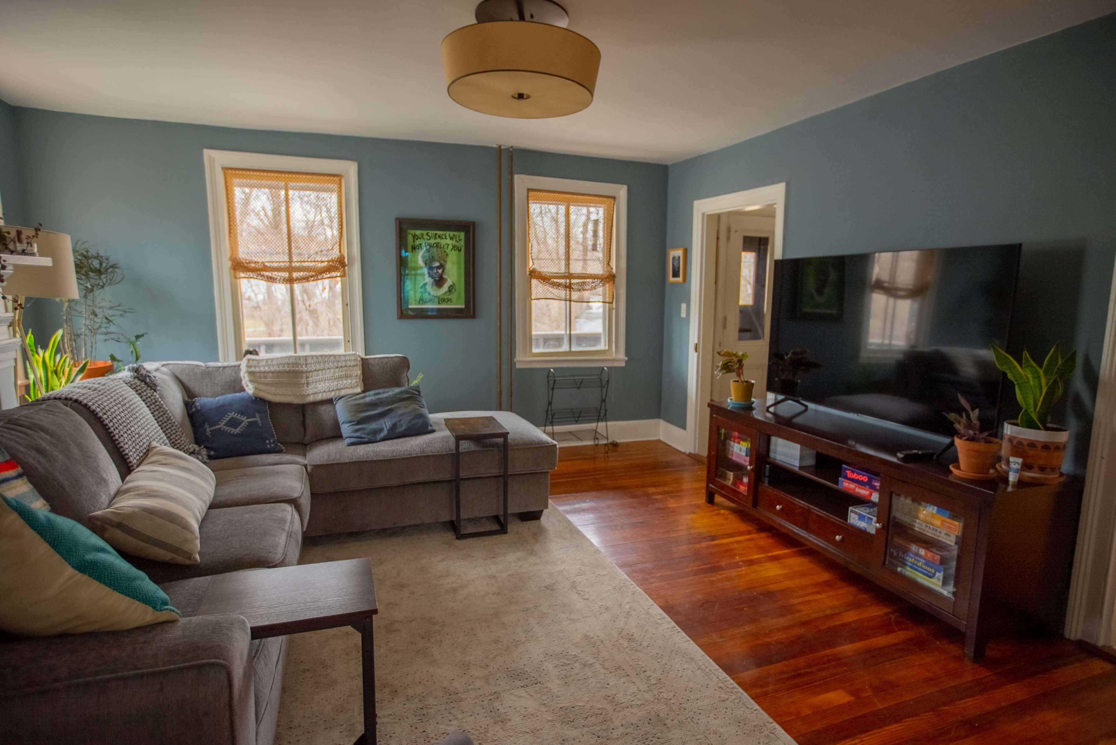A cozy living room features a gray sectional sofa, a large television on a wooden stand, and windows with light-filtering shades.