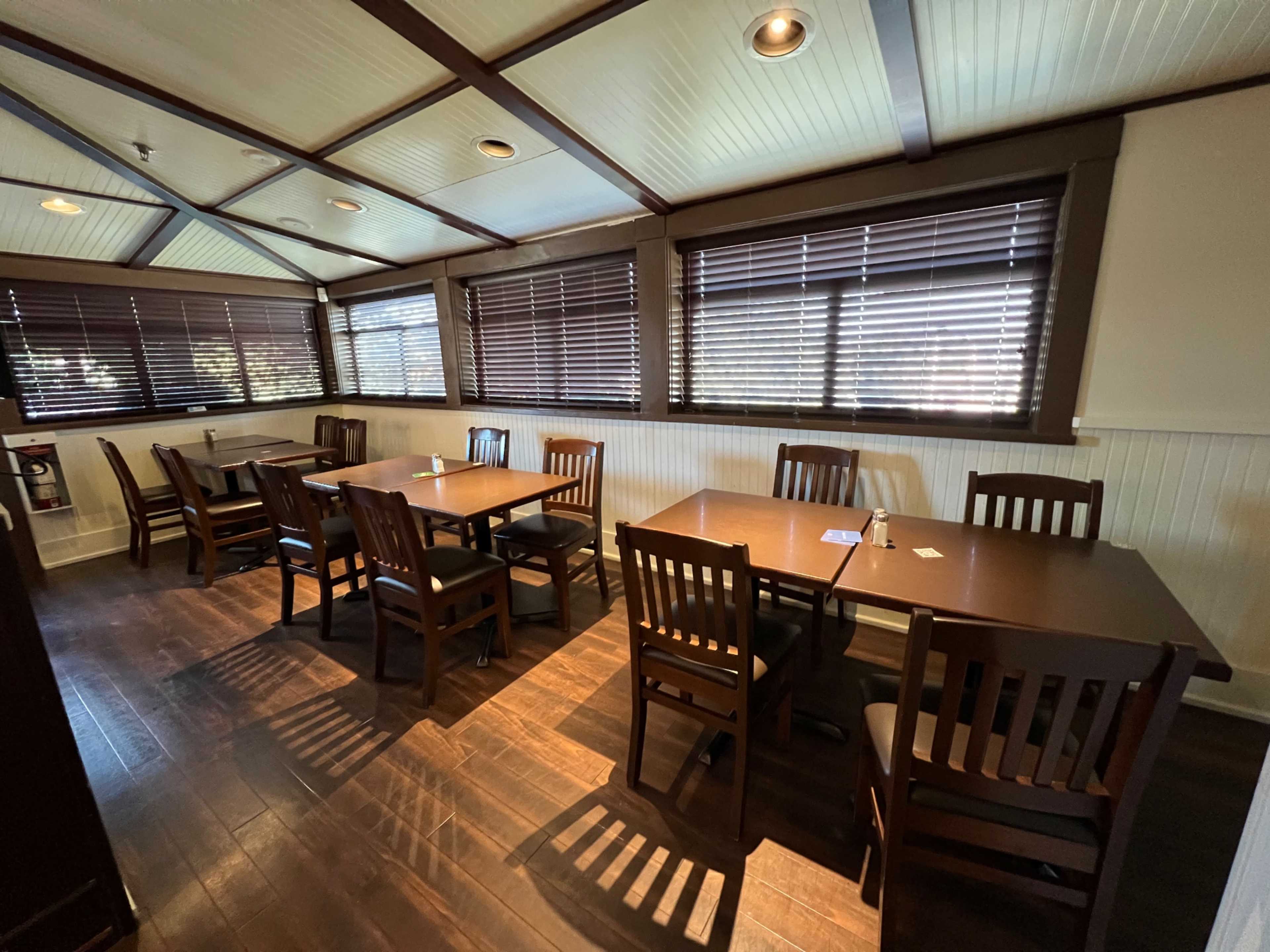An empty dining area with wooden tables and chairs, featuring blinds on the windows and a dark wood floor.
