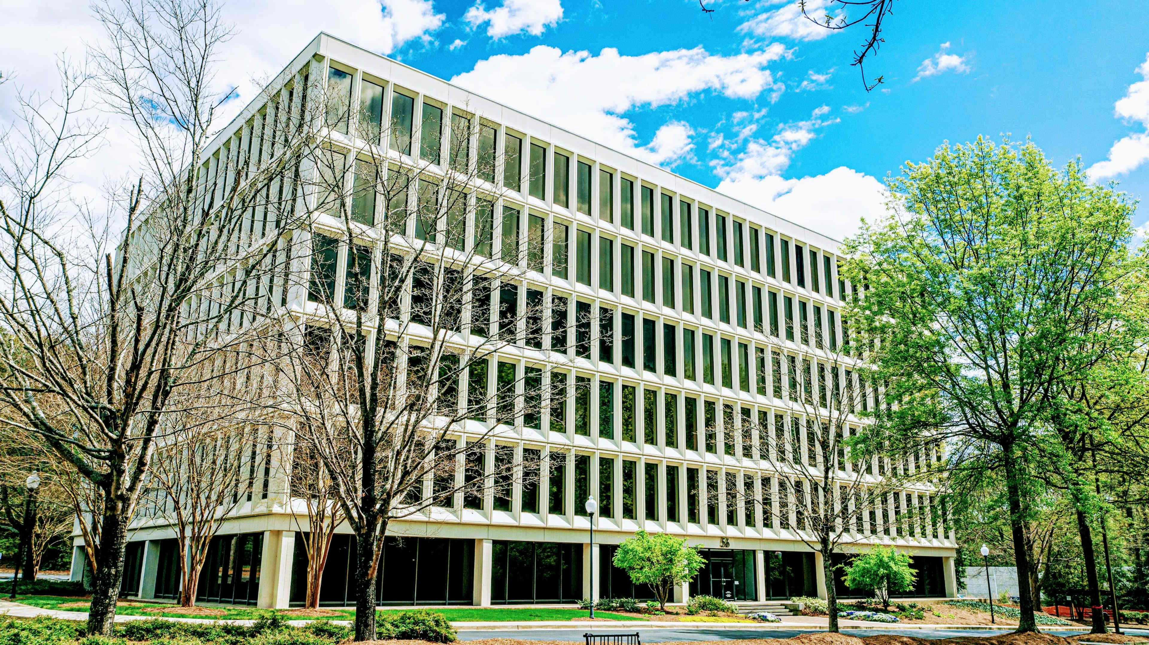A modern, multi-story office building with numerous large windows is surrounded by bare trees and young green foliage under a blue sky.