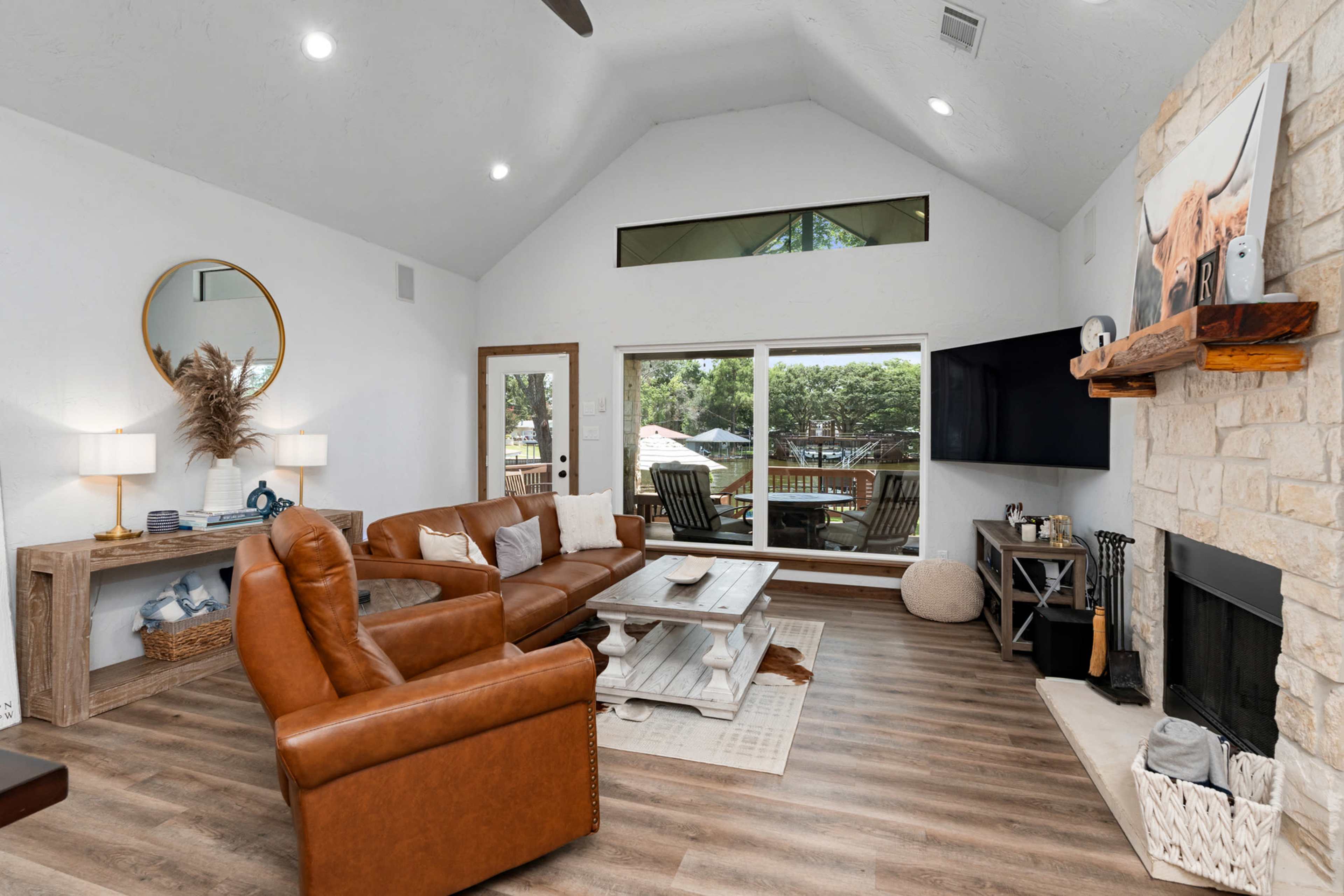 The living room features a brown leather sectional sofa, a wooden coffee table, and large windows that overlook an outdoor seating area.