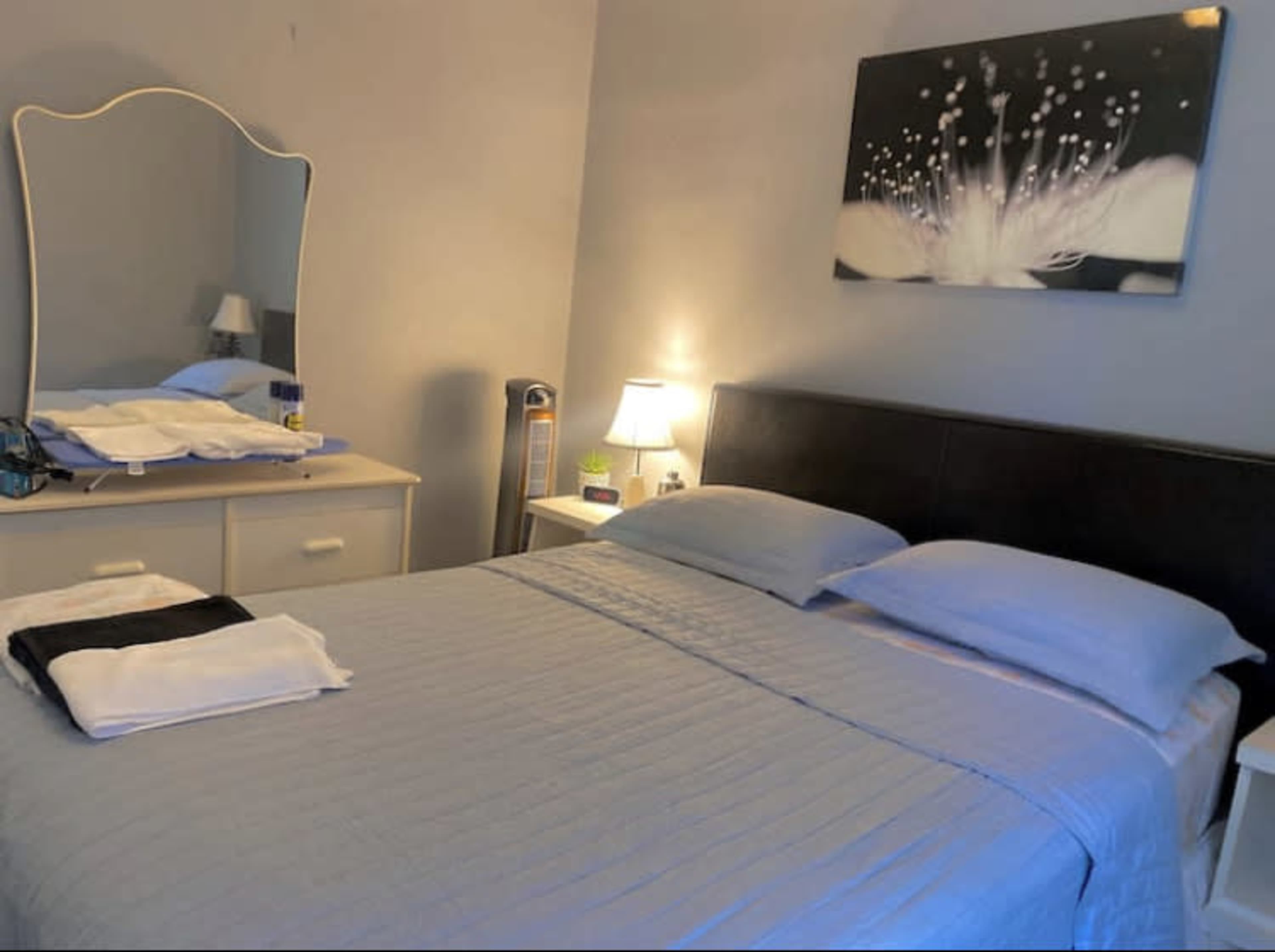 A bedroom with a neatly made bed, a dresser with a mirror, a bedside lamp, and a decorative wall picture of a flower.