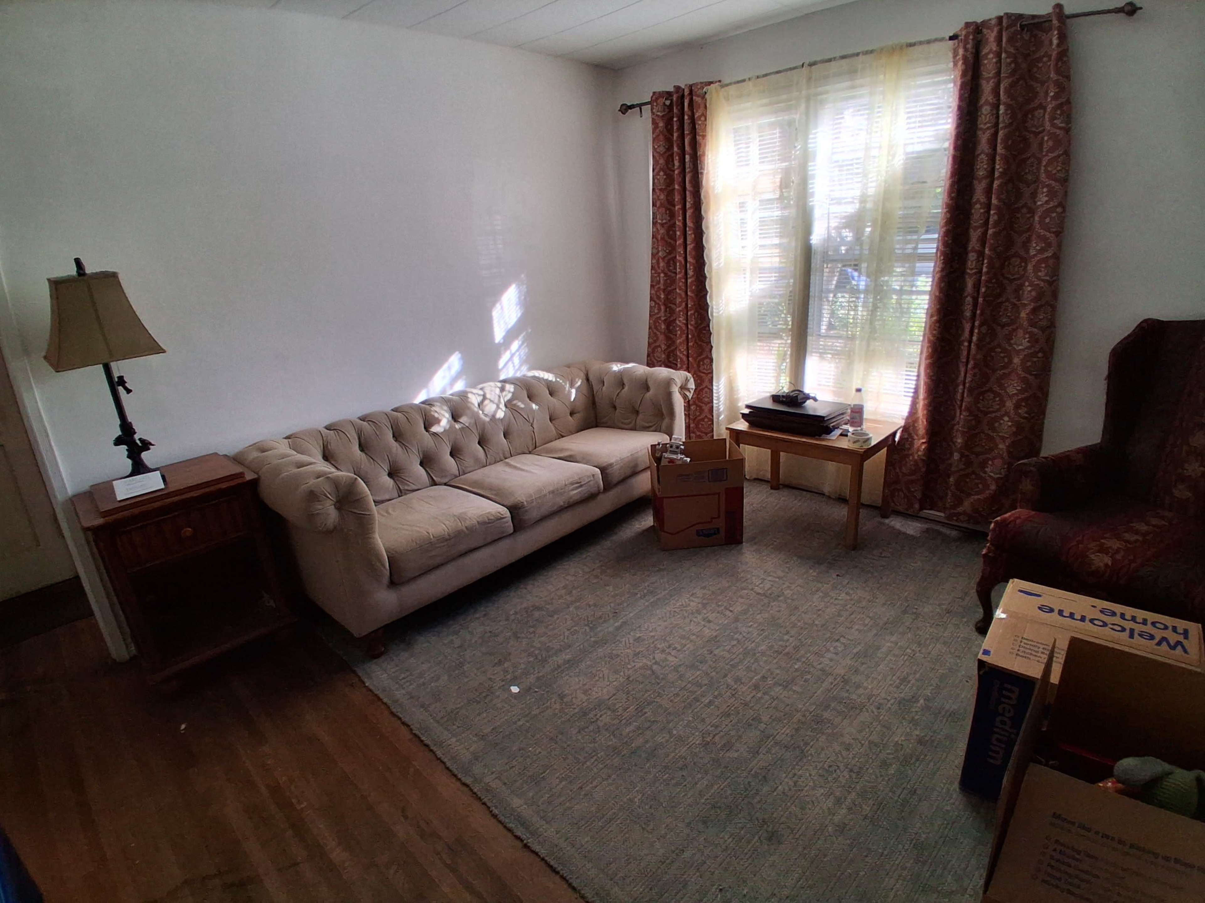 The image shows a partially furnished living room with a beige tufted sofa, a small wooden table beside it, a patterned chair in the corner, and several cardboard boxes on the floor.