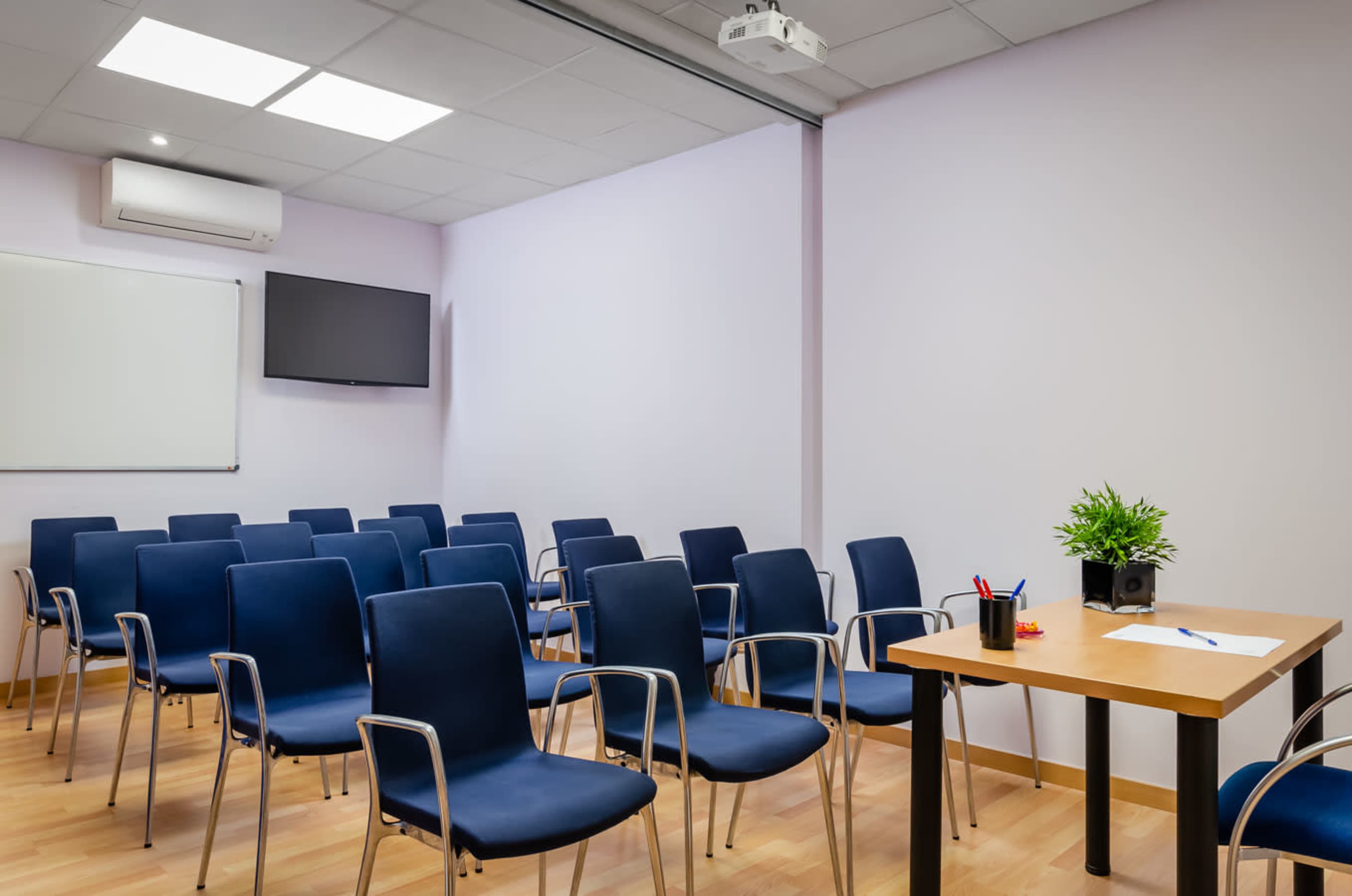 A conference room with several blue chairs arranged in rows facing a small table and a projector screen.