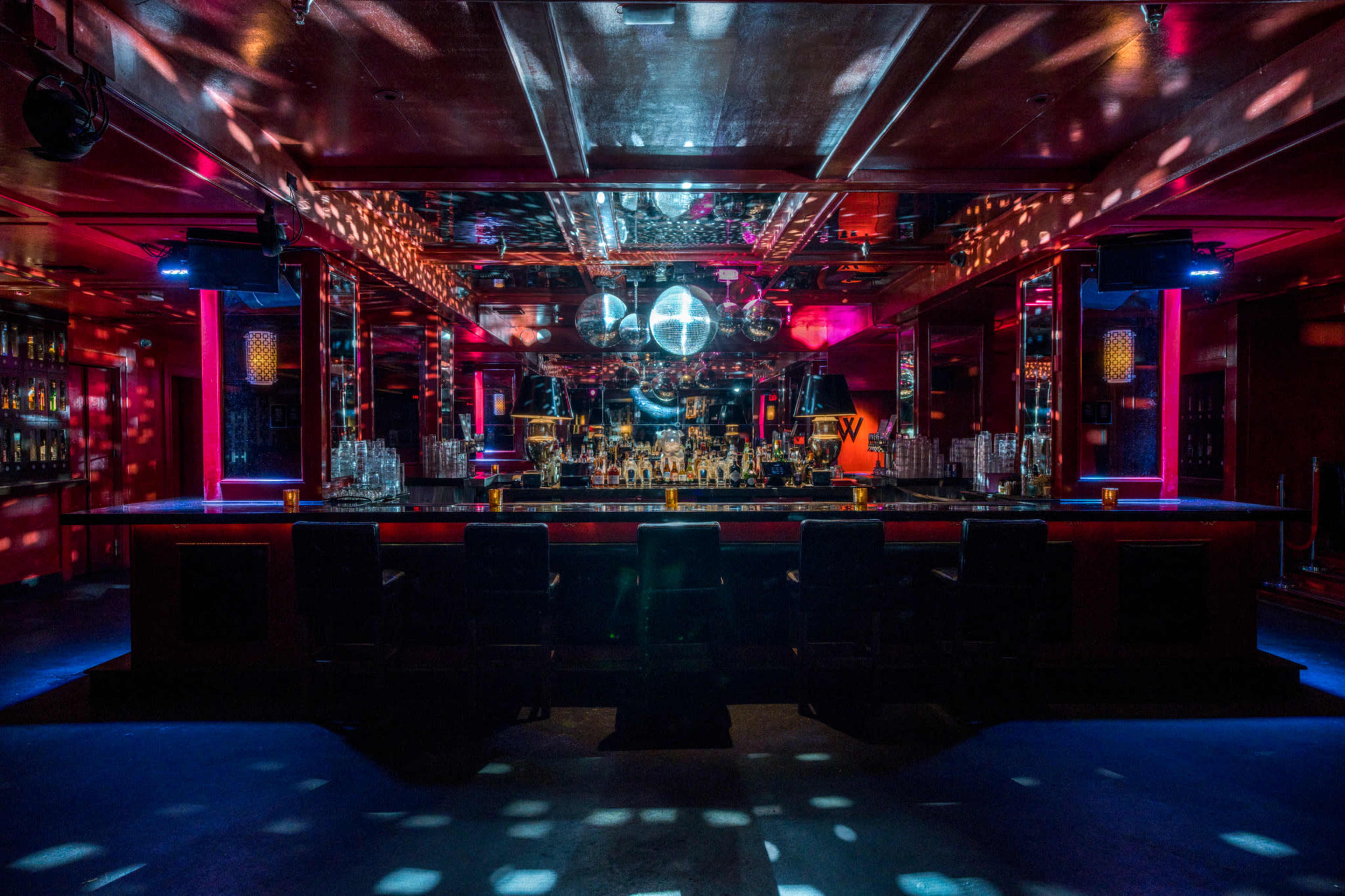 A dimly lit bar with a spacious counter, disco balls reflecting colorful lights, and a row of stools.