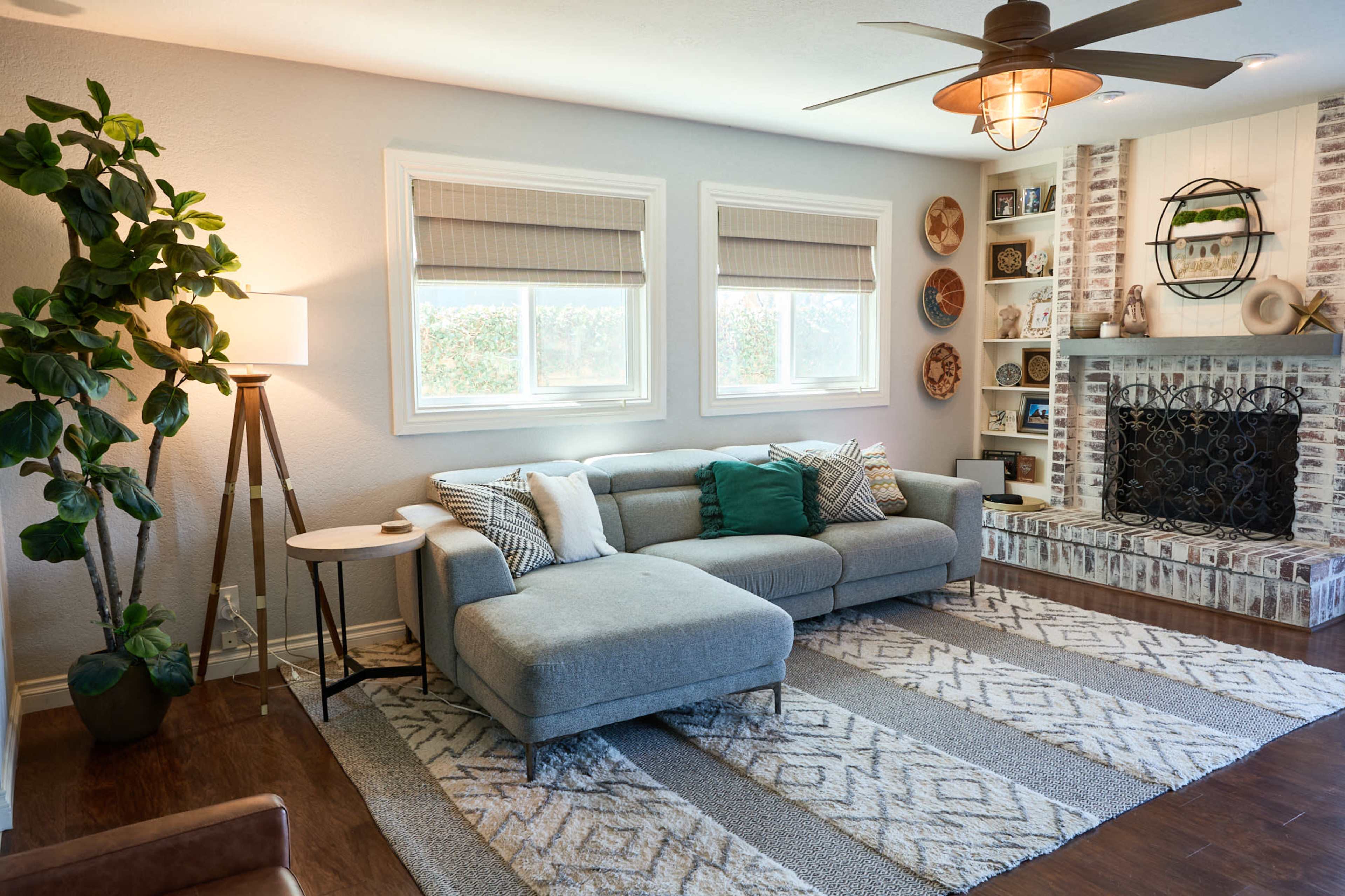 The living room features a gray sectional sofa, a decorative rug, and a brick fireplace with shelves, illuminated by a ceiling fan and natural light from two windows.
