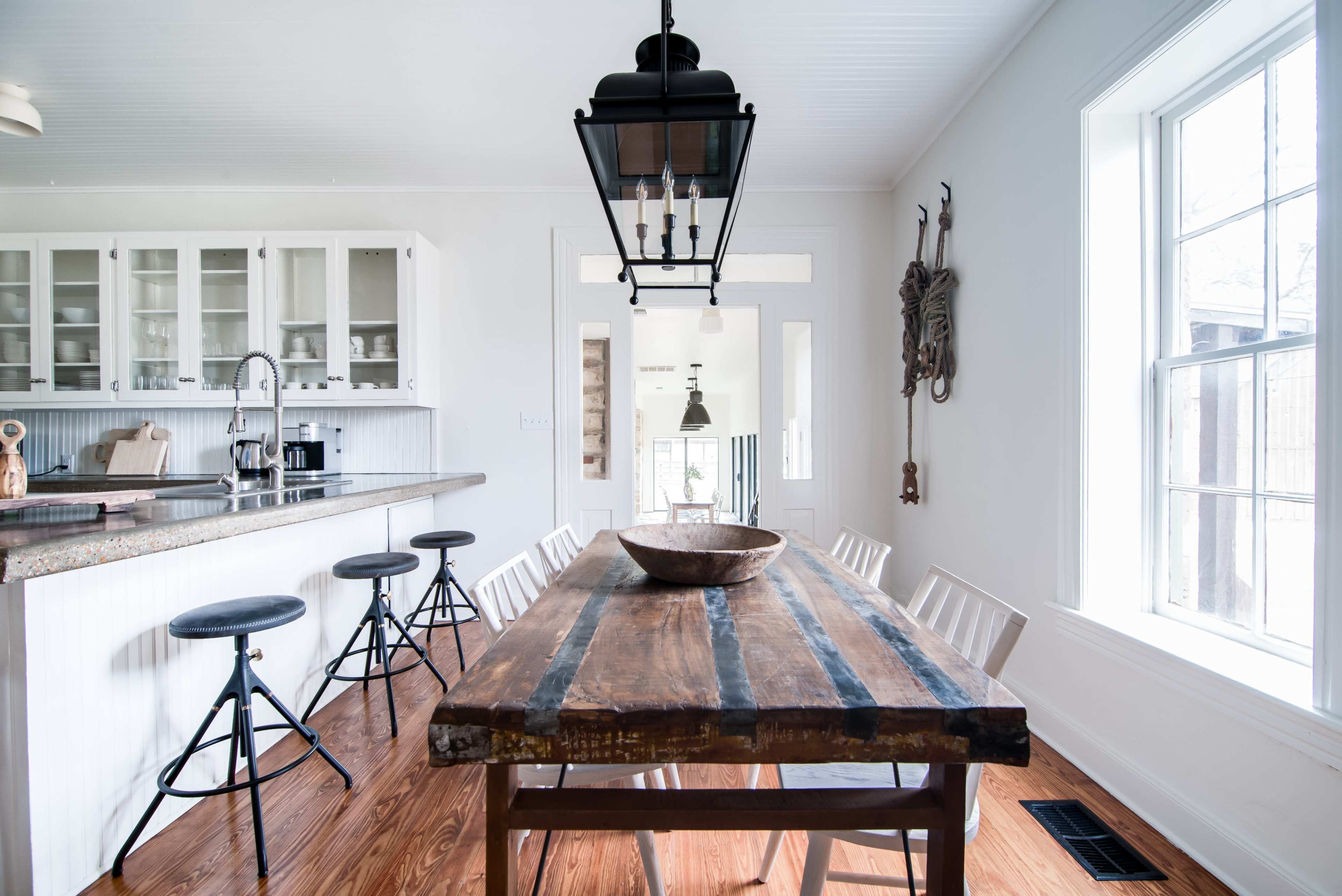 A modern kitchen features a large wooden dining table surrounded by white chairs, with a countertop and bar stools, and large windows allowing natural light.