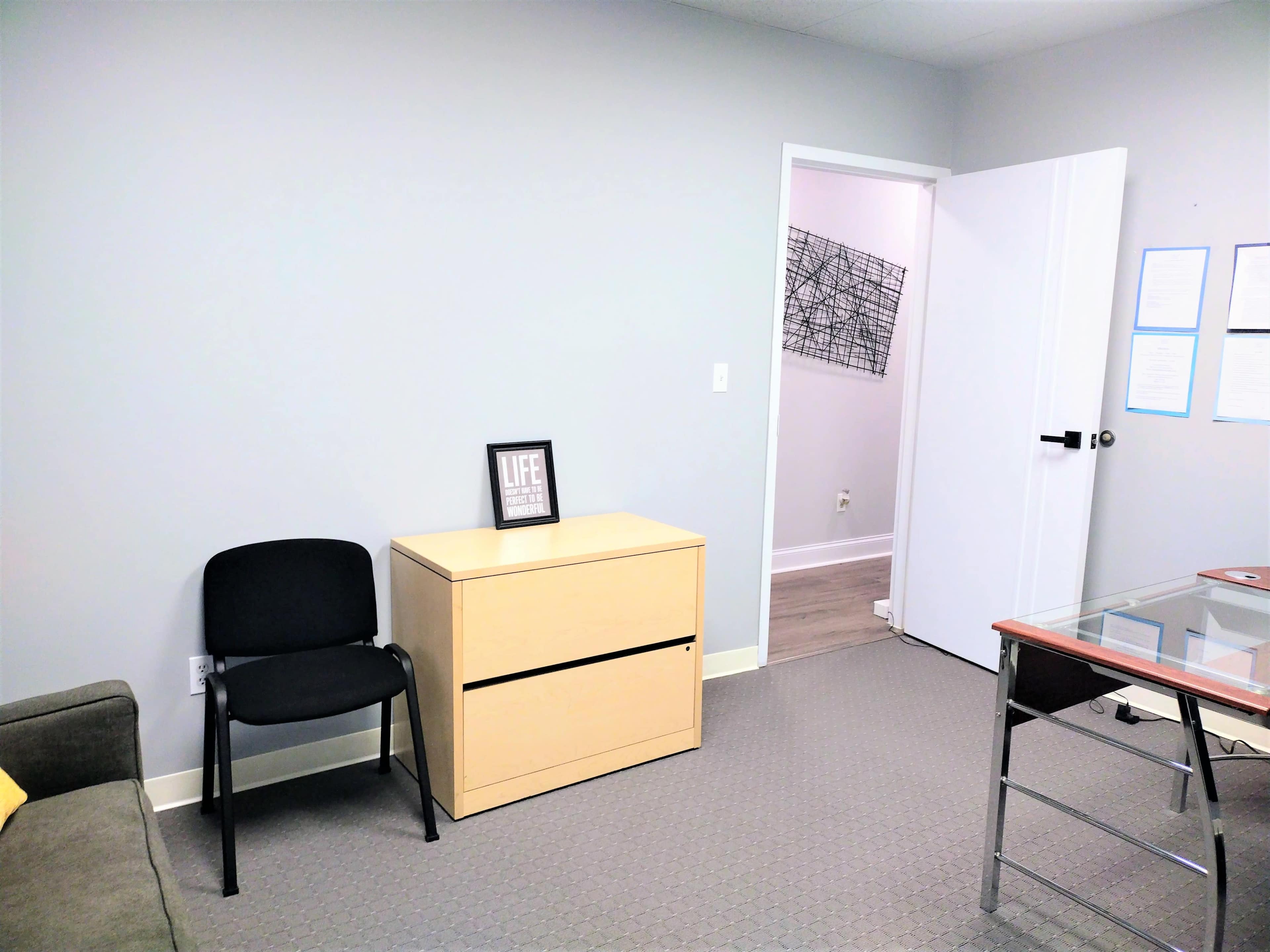 A minimalistic office space featuring a black chair, a wooden filing cabinet, and a glass-top desk, with a doorway leading to another room.