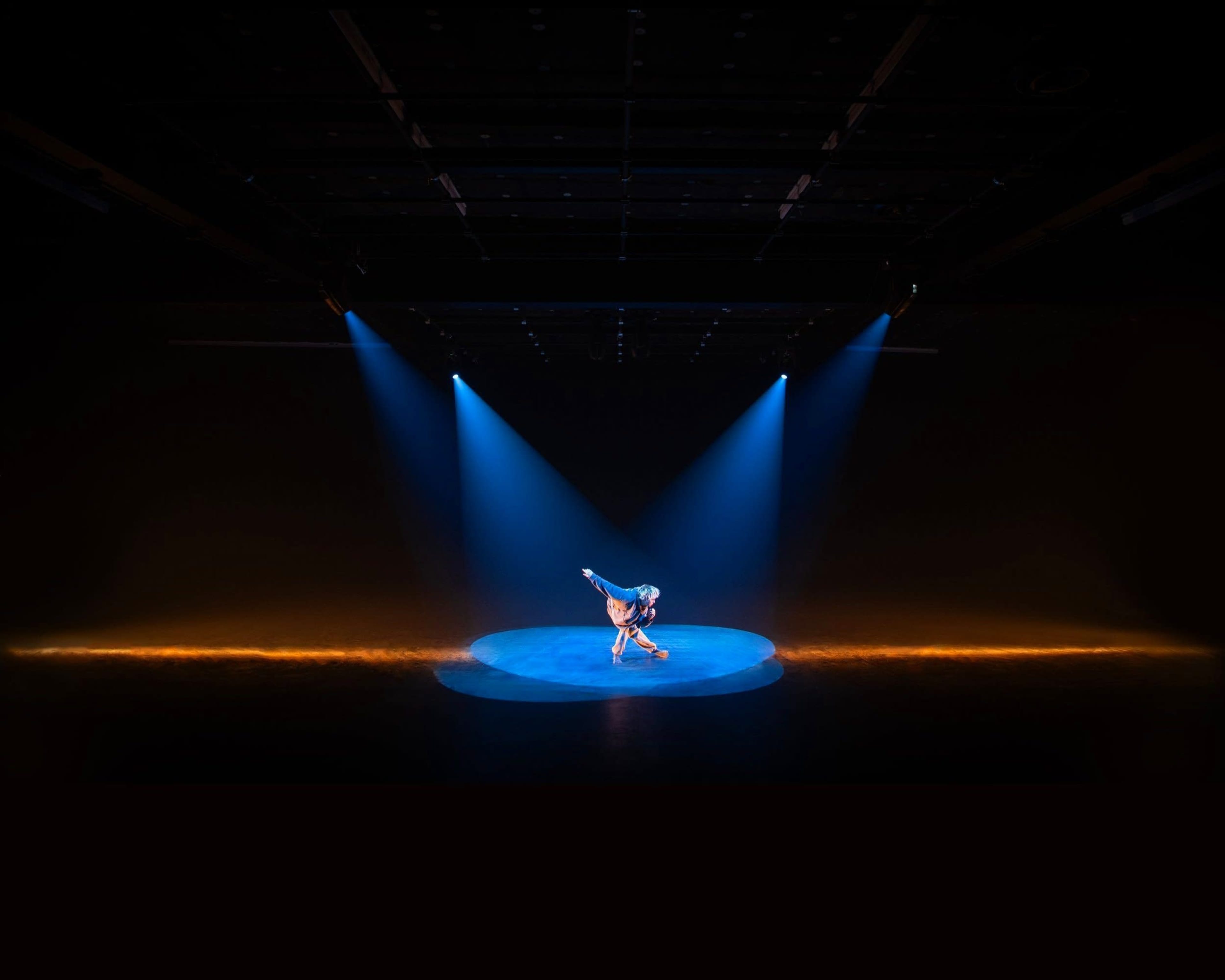 A performer strikes a pose on a circular stage illuminated by blue spotlight beams against a dark background.