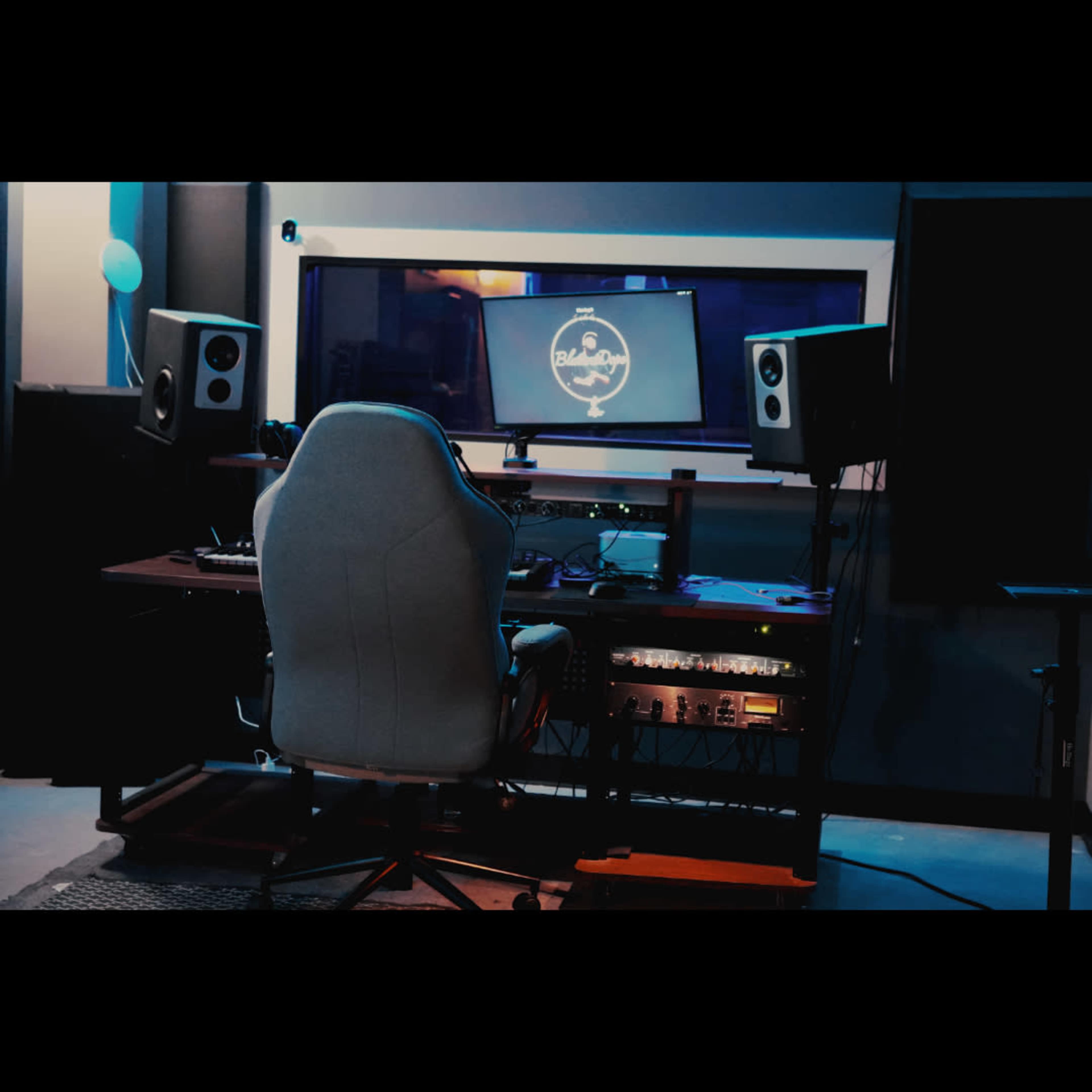 The image shows a modern music studio setup featuring a computer monitor, audio speakers, and a central desk with a high-backed chair facing the workspace.
