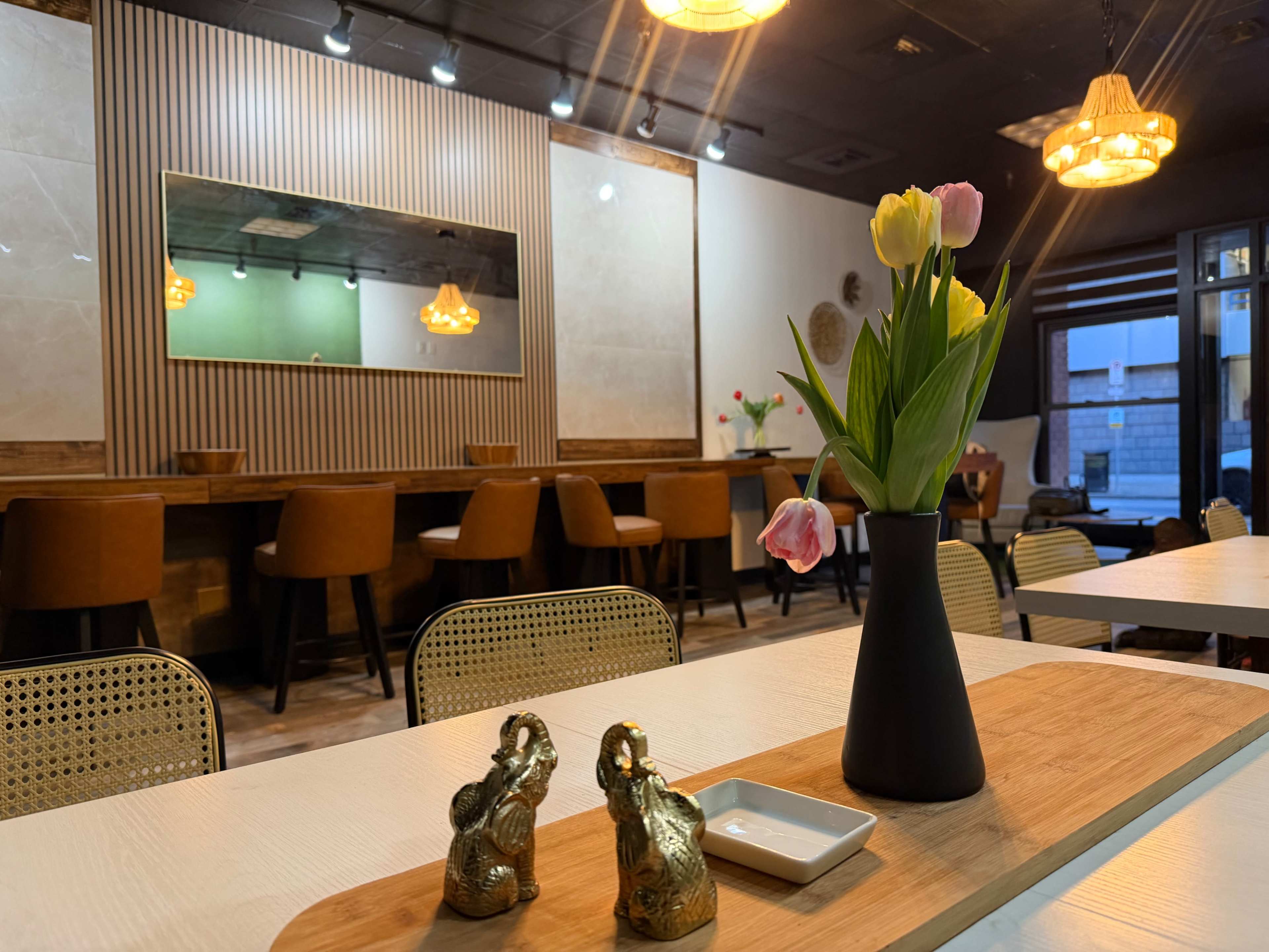 A modern café interior features a long wooden table with a vase of tulips, gold elephant decorations, and a bar area with high-backed chairs.
