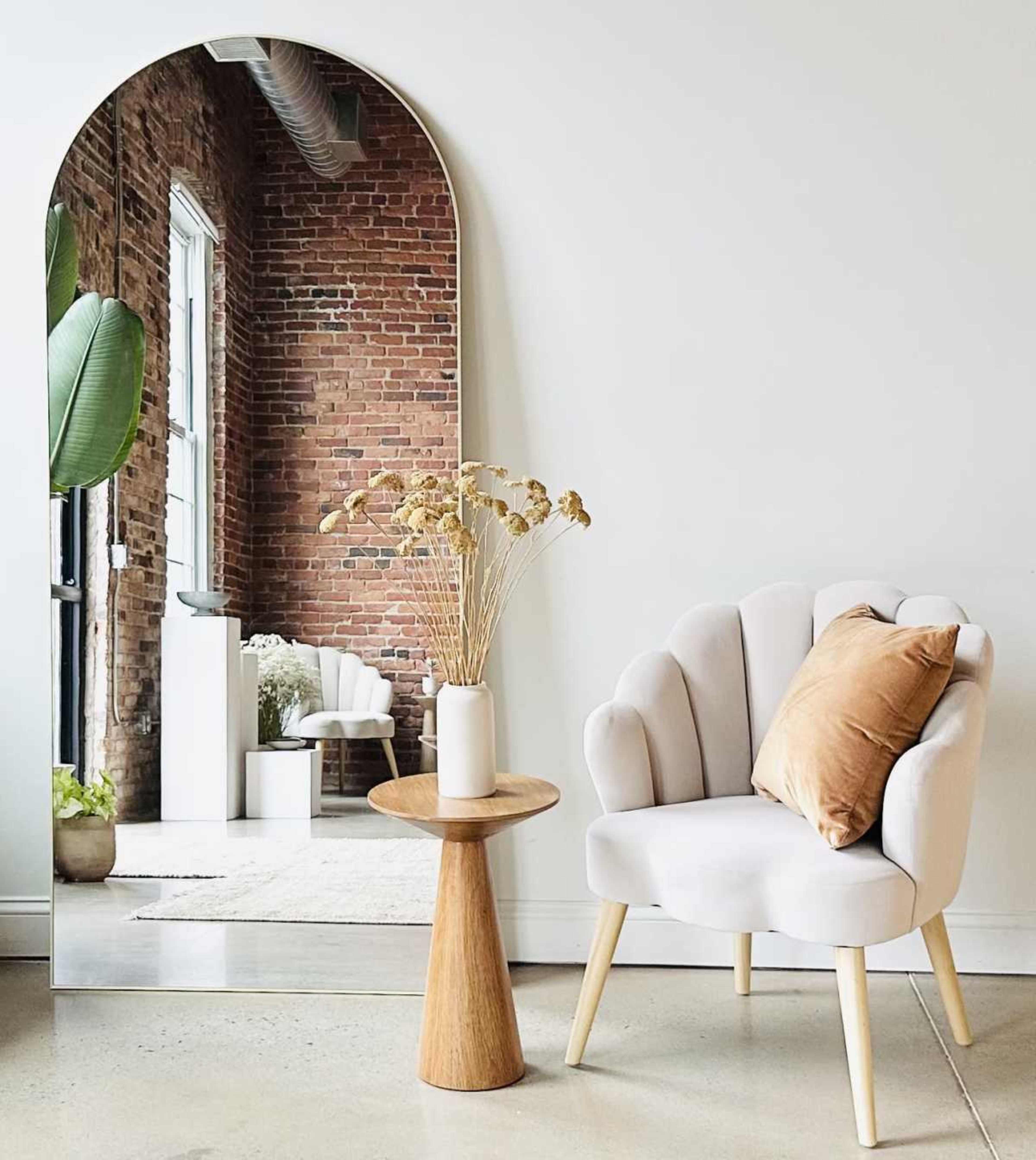 The image features a modern chair beside a wooden side table, with a tall arched mirror reflecting a brick wall and indoor plant in a minimalist interior space.
