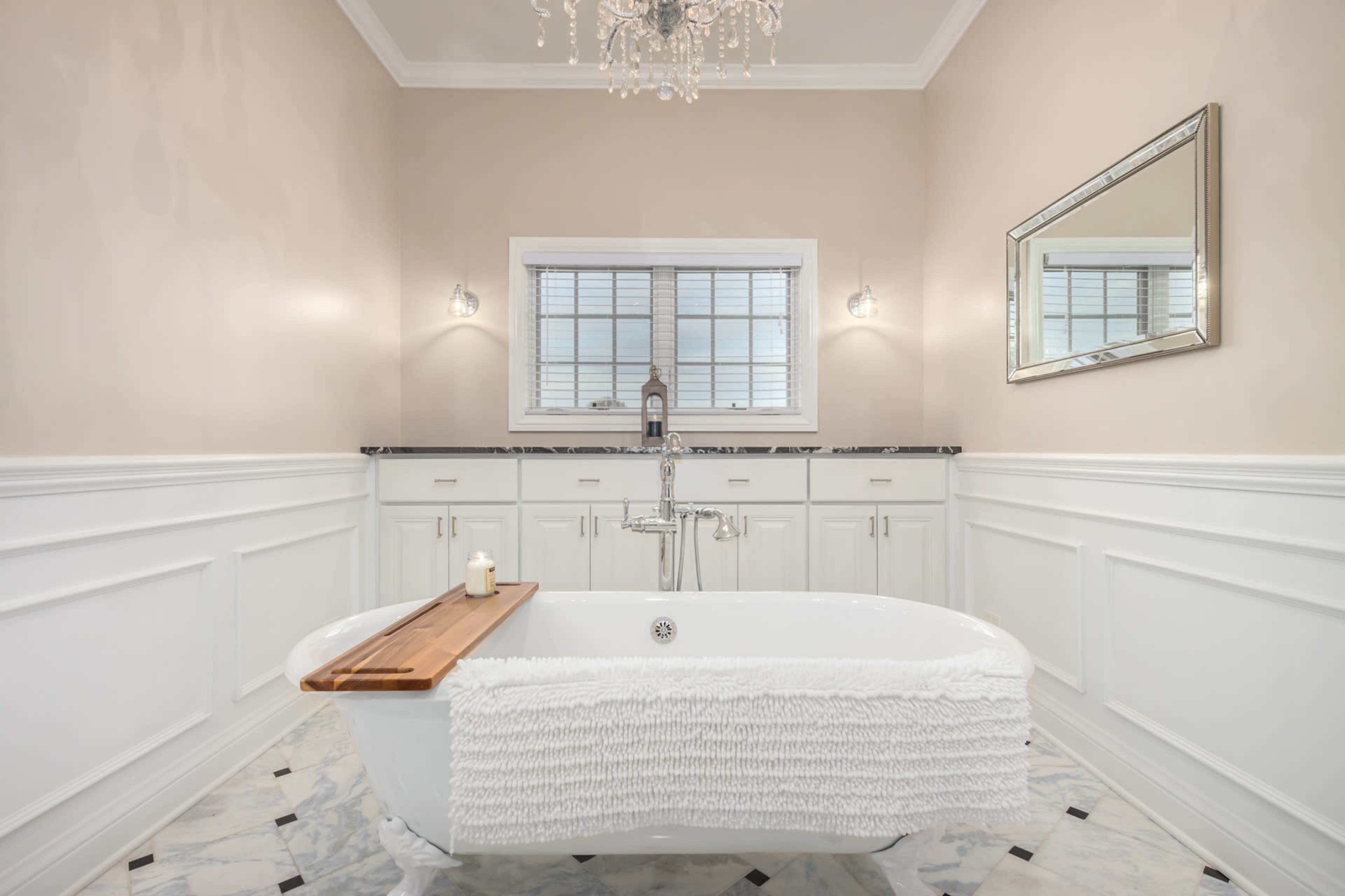 A spacious bathroom featuring a freestanding bathtub, a marble floor, and a window with a decorative mirror above a vanity unit.