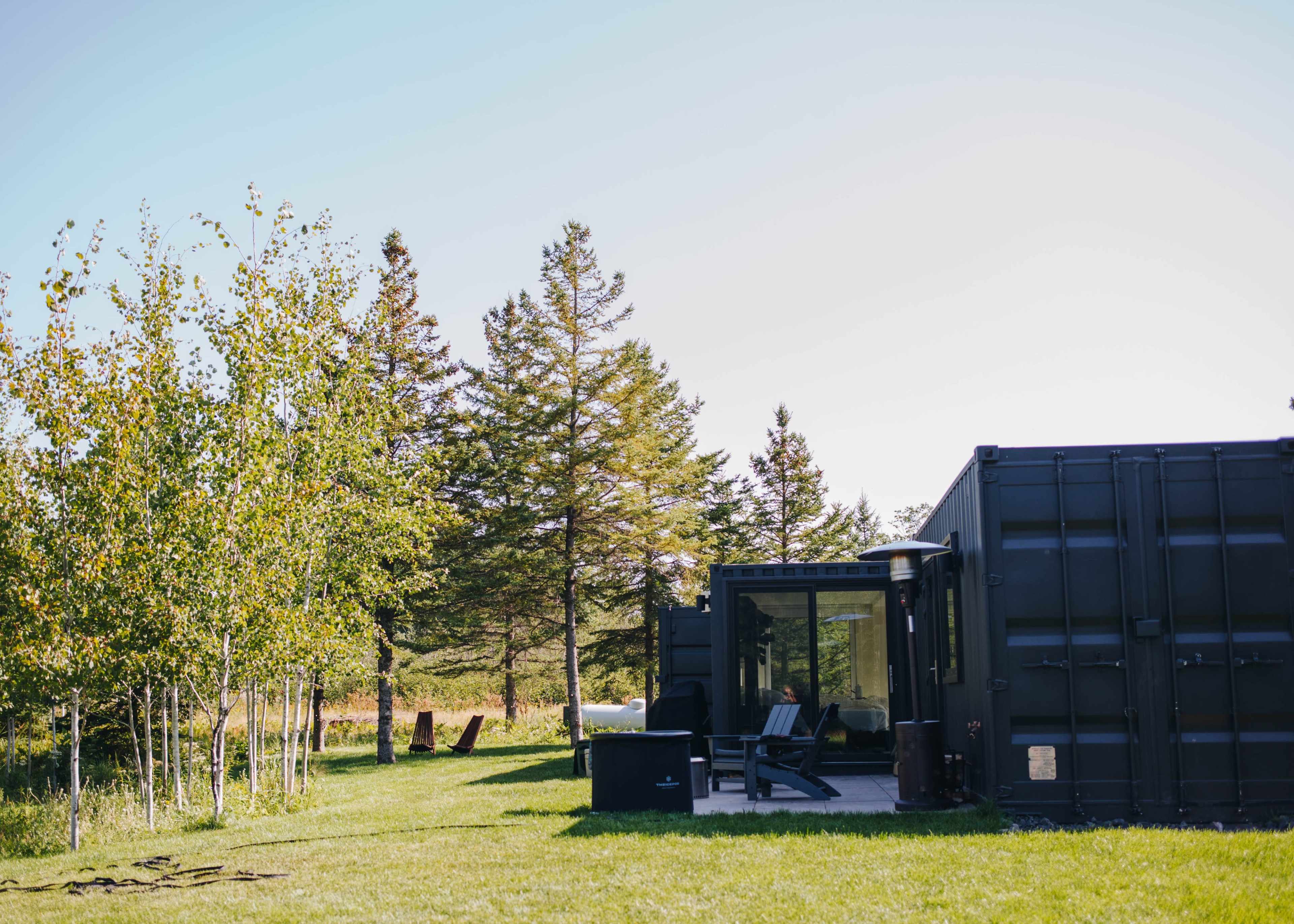 A modern black container home is set against a backdrop of green grass and trees under a clear sky.